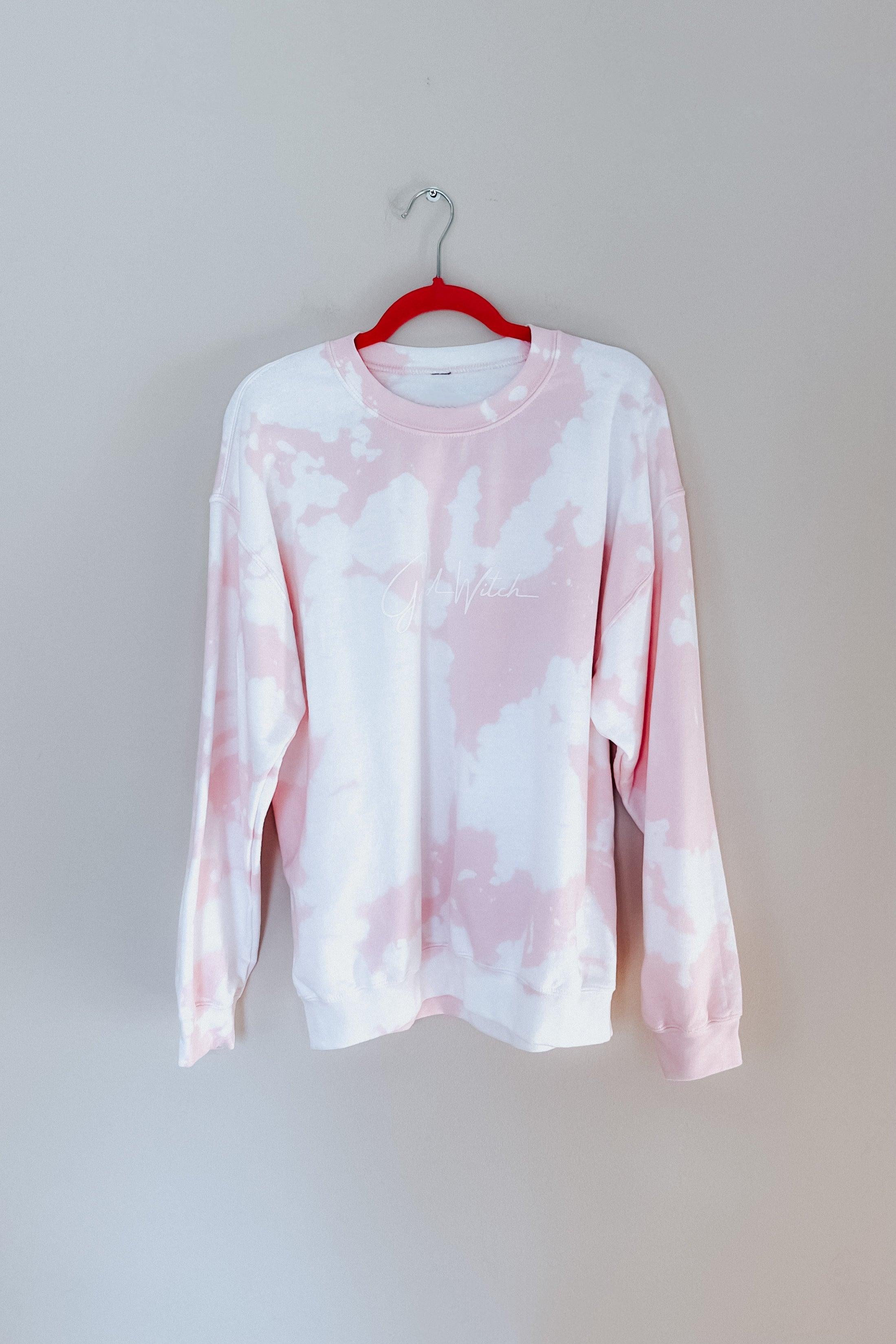 Pastel pink and white tie-dye pullover with subtle script design hanging on red hanger against plain wall