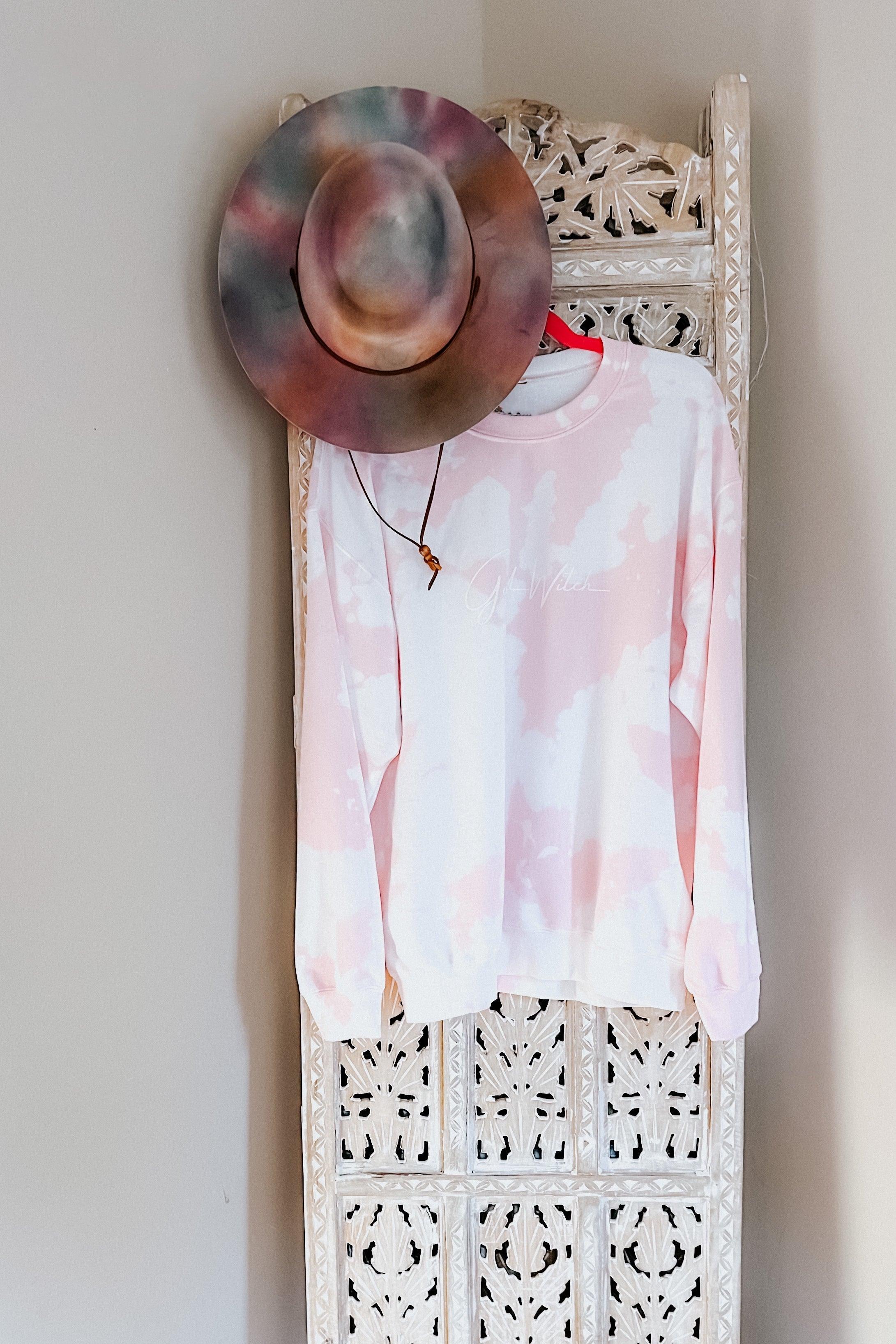 Pastel pink and white bleached pullover with subtle script hanging on a decorative carved wooden panel with a multicolor hat above