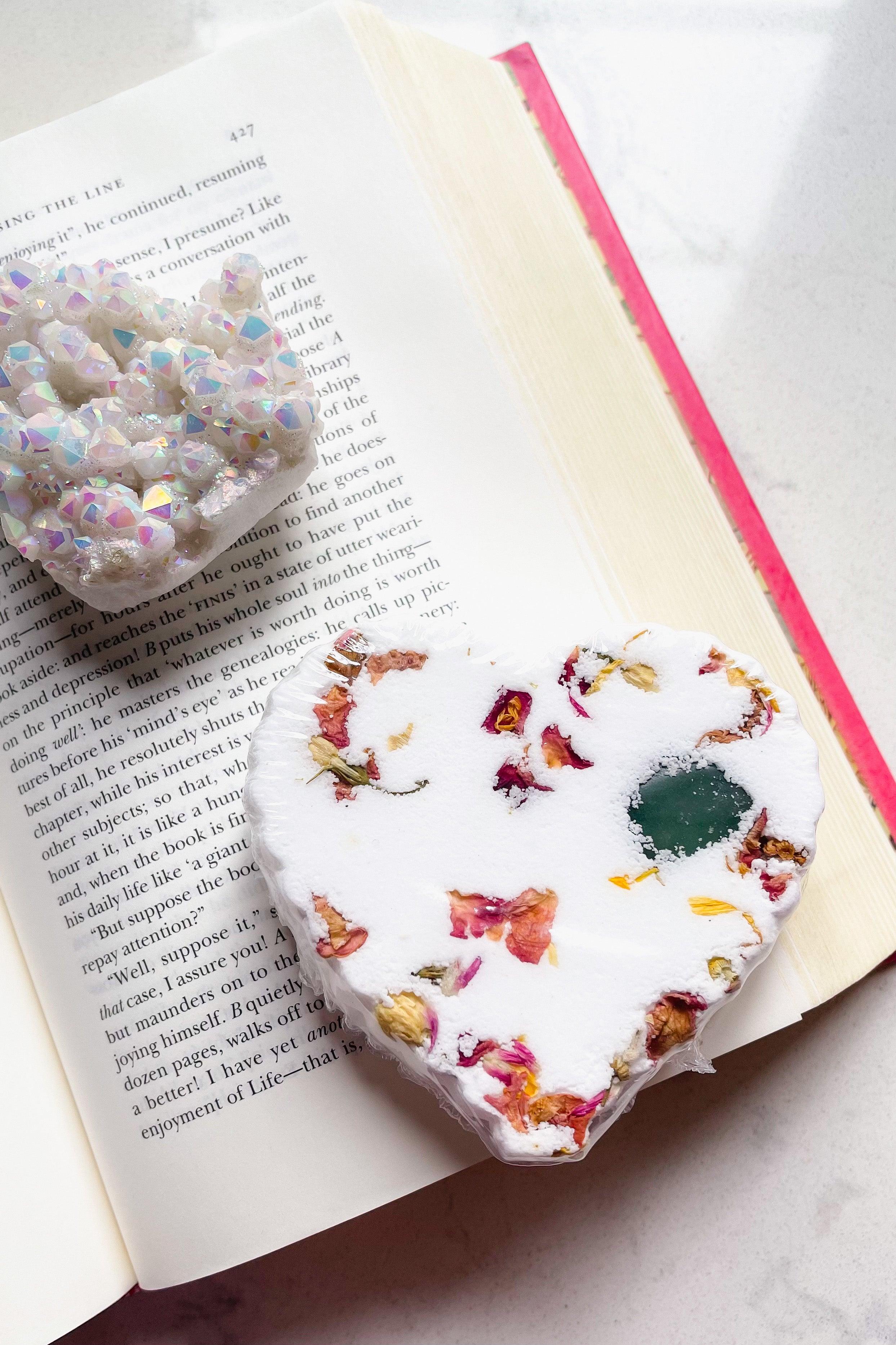 White heart-shaped bath bomb with embedded dried flower petals and green crystal detail on open book pages