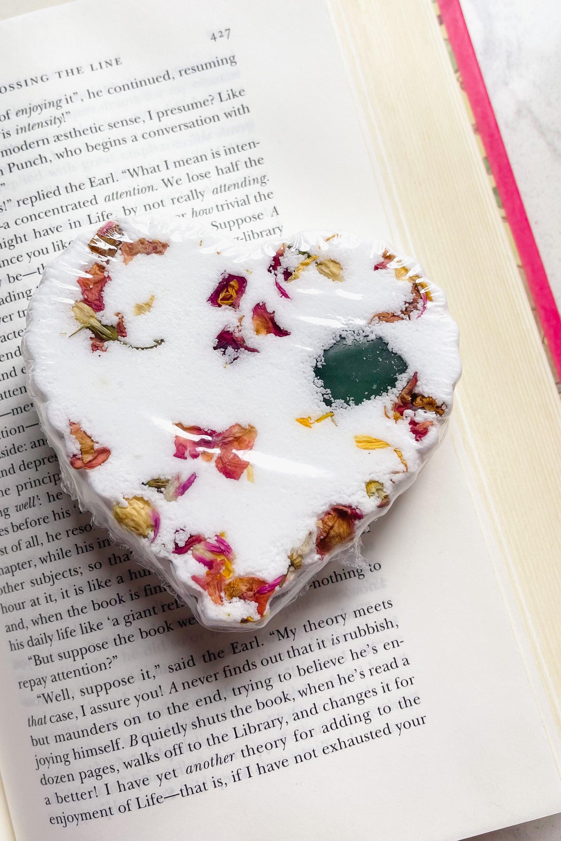 White heart-shaped bath bomb with dried flower petals and a green crystal centerpiece resting on an open book page
