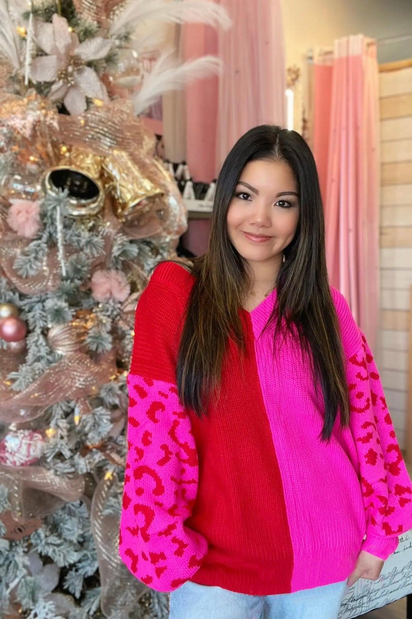 Woman wearing plush pink and red color block sweater with leopard print sleeves standing by decorated Christmas tree