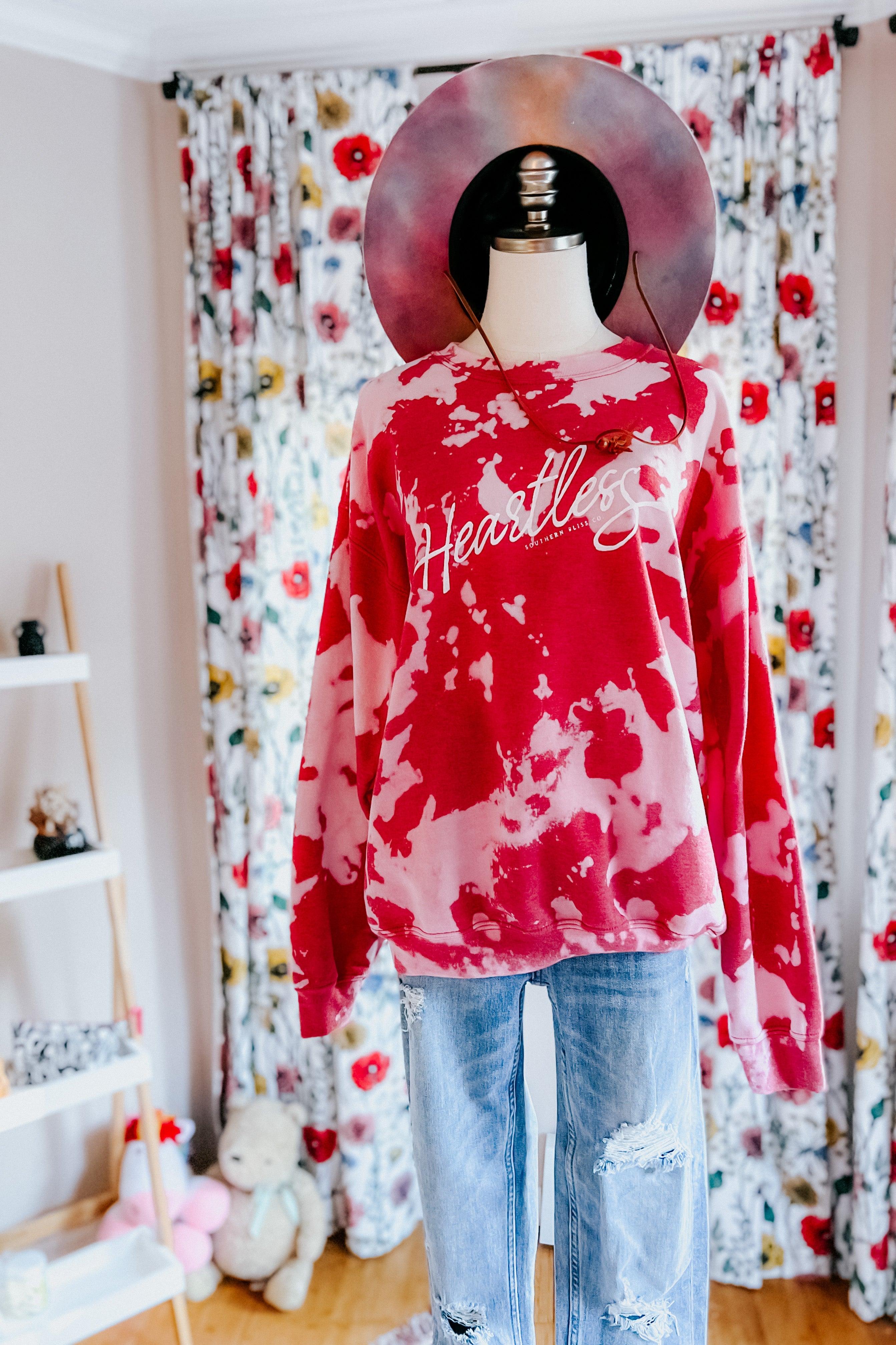 Red bleached pullover with white "Heartless" script logo displayed on mannequin styled with distressed blue jeans and wide-brim hat