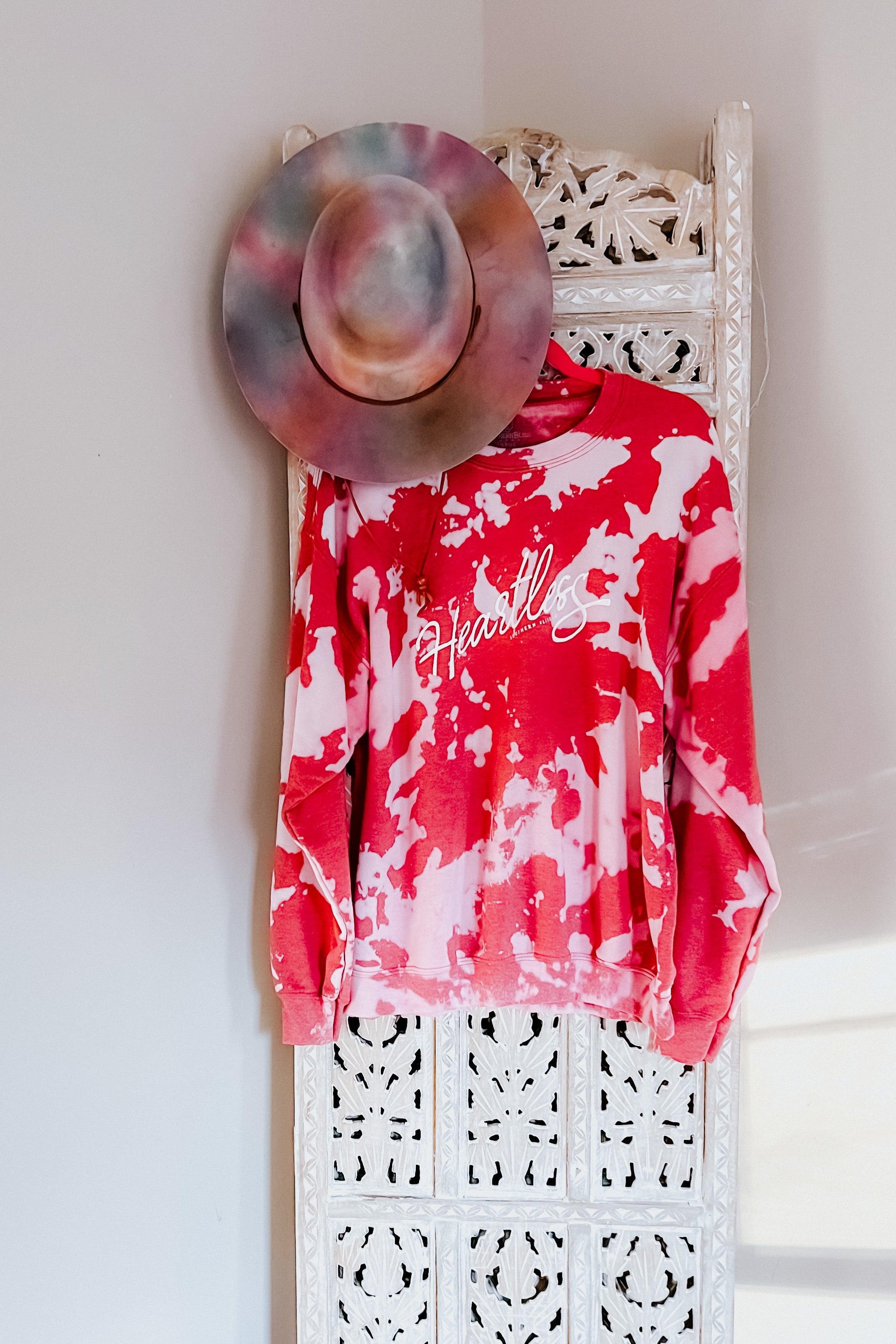 Red tie-dye bleached pullover with white "Heartless" script logo hanging on carved white wooden panel with multicolor hat