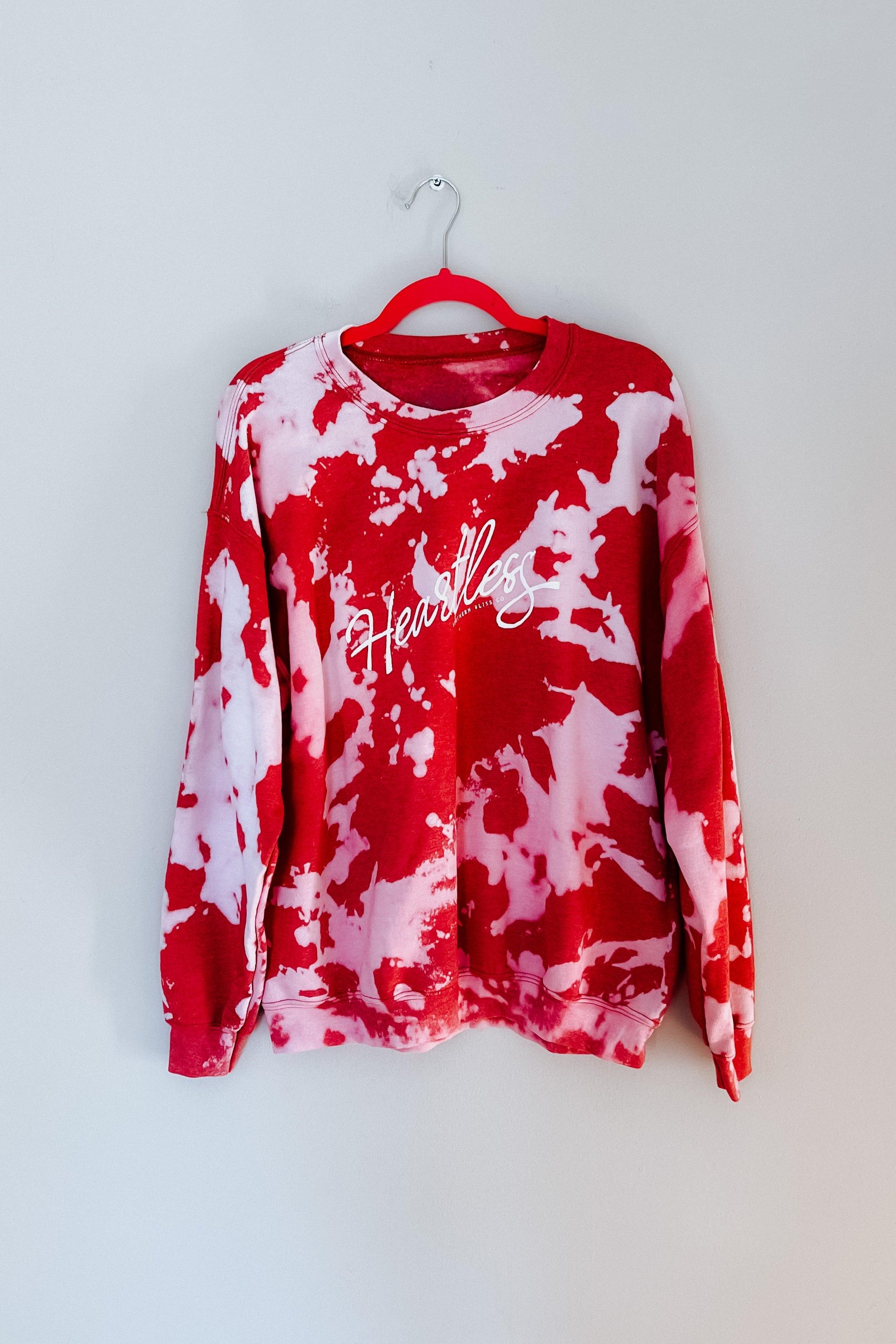 Red bleached pullover with white "Heartless" script logo hanging on red hanger against plain background