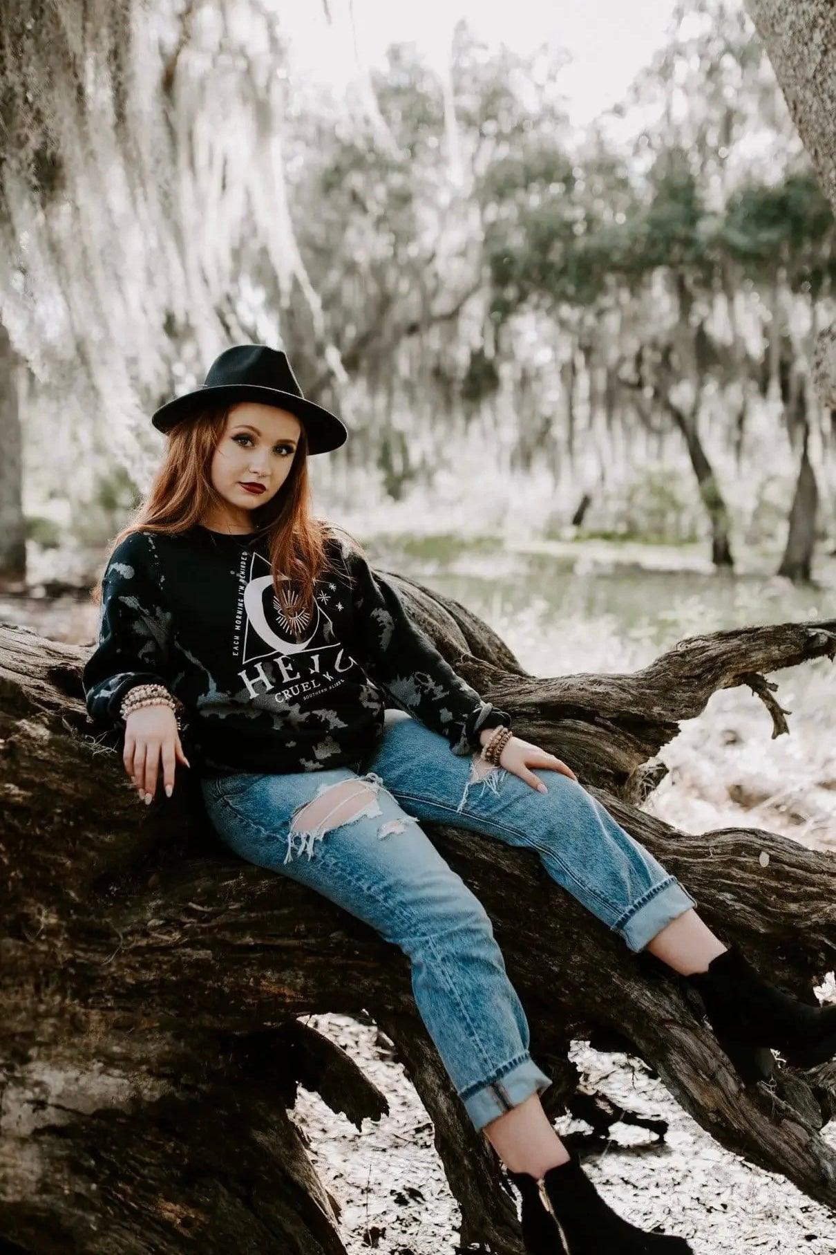 Woman seated outdoors on large tree limb wearing black Hello Cruel World bleached sweatshirt, distressed jeans, and wide-brim hat
