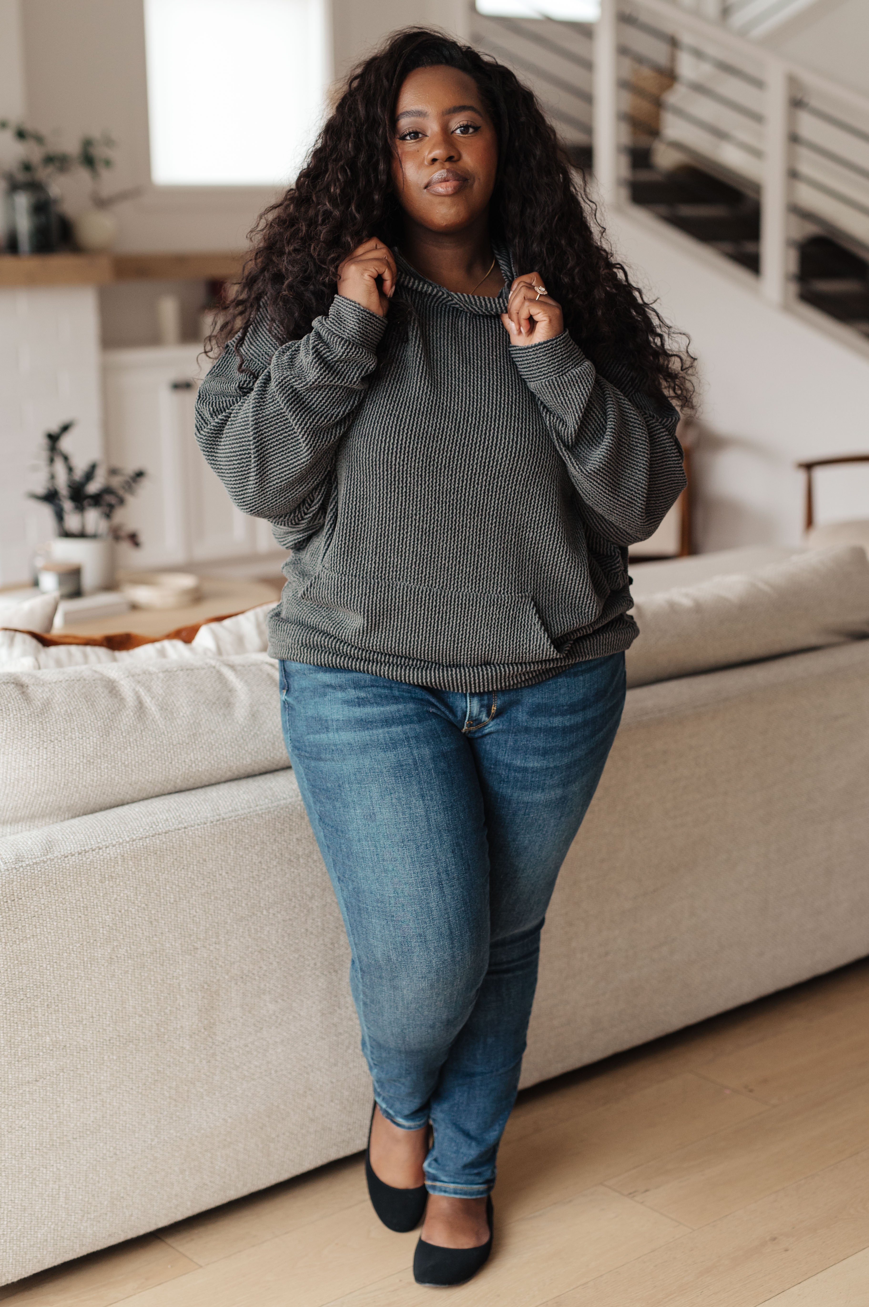 A plus size woman wearing a grey rib knit hoodie over blue jeans and black ballet flats against an indoor setting