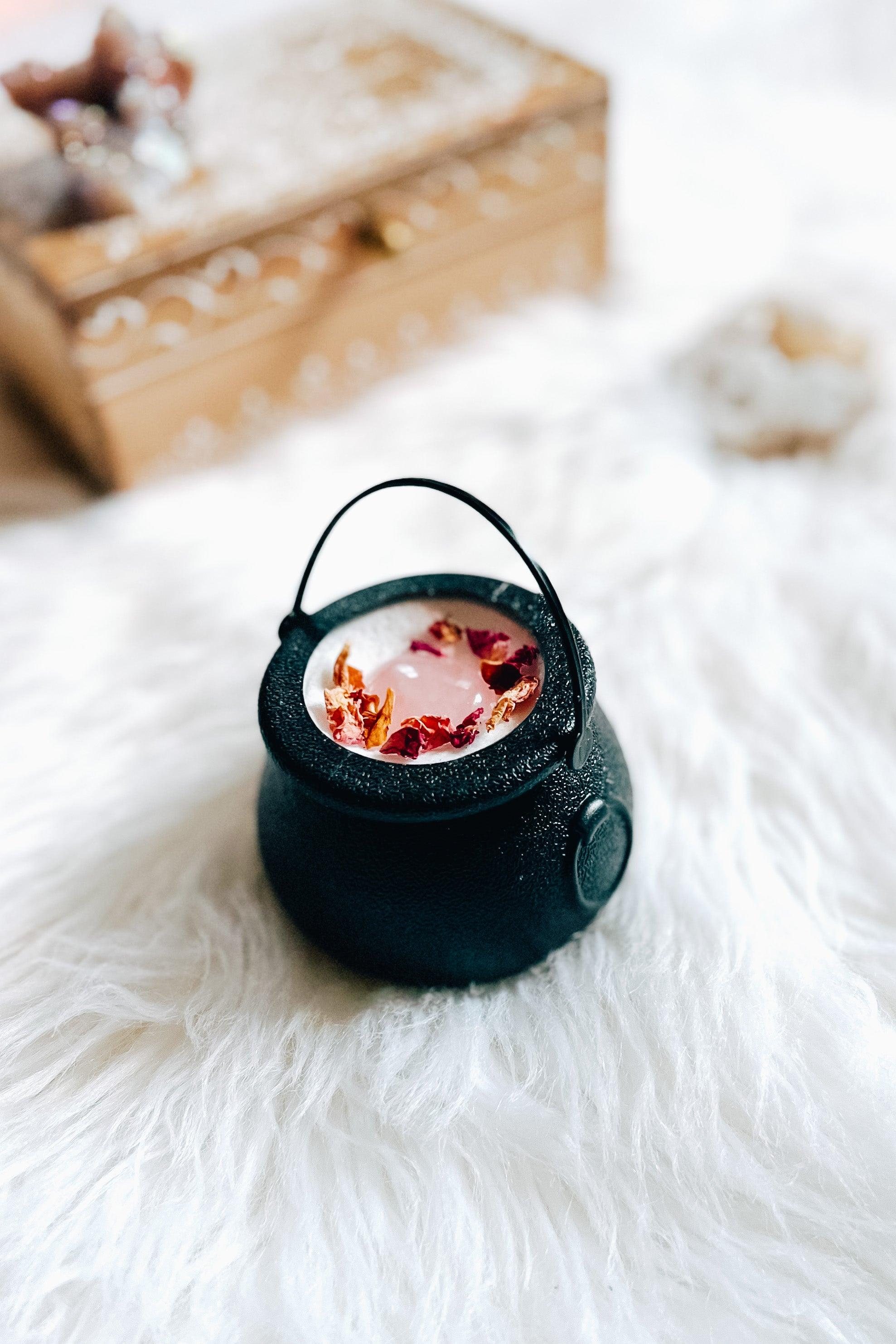 Black cauldron bath bomb with pink heart shaped rose quartz and dried rose petals resting on fluffy white fur with wooden box blurred behind