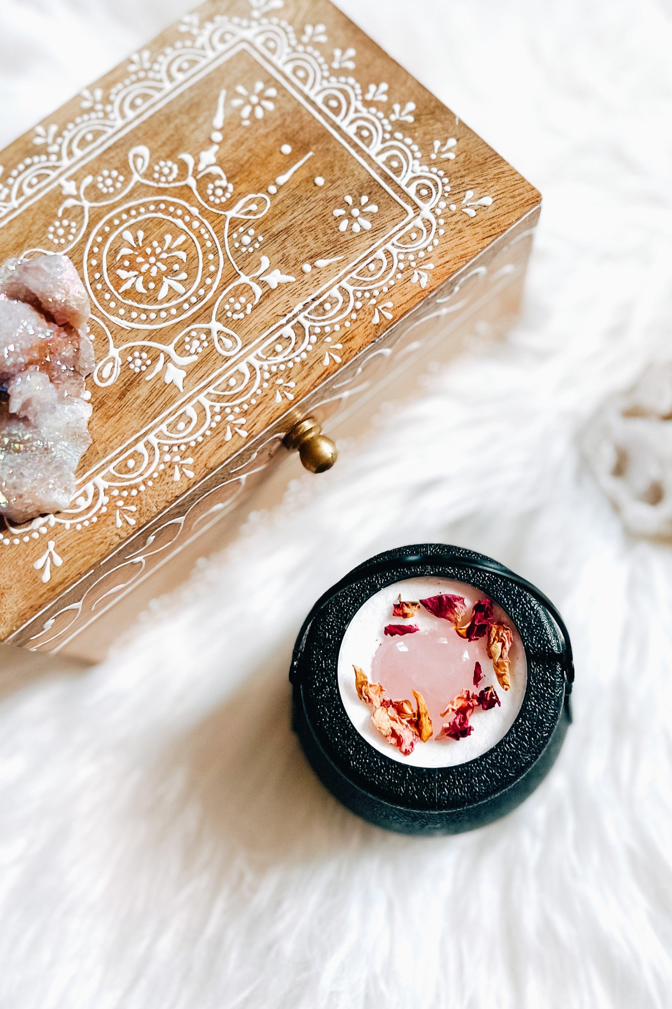 Black cauldron bath bomb with pink heart-shaped crystal and dried rose petals next to wooden box with white painted patterns