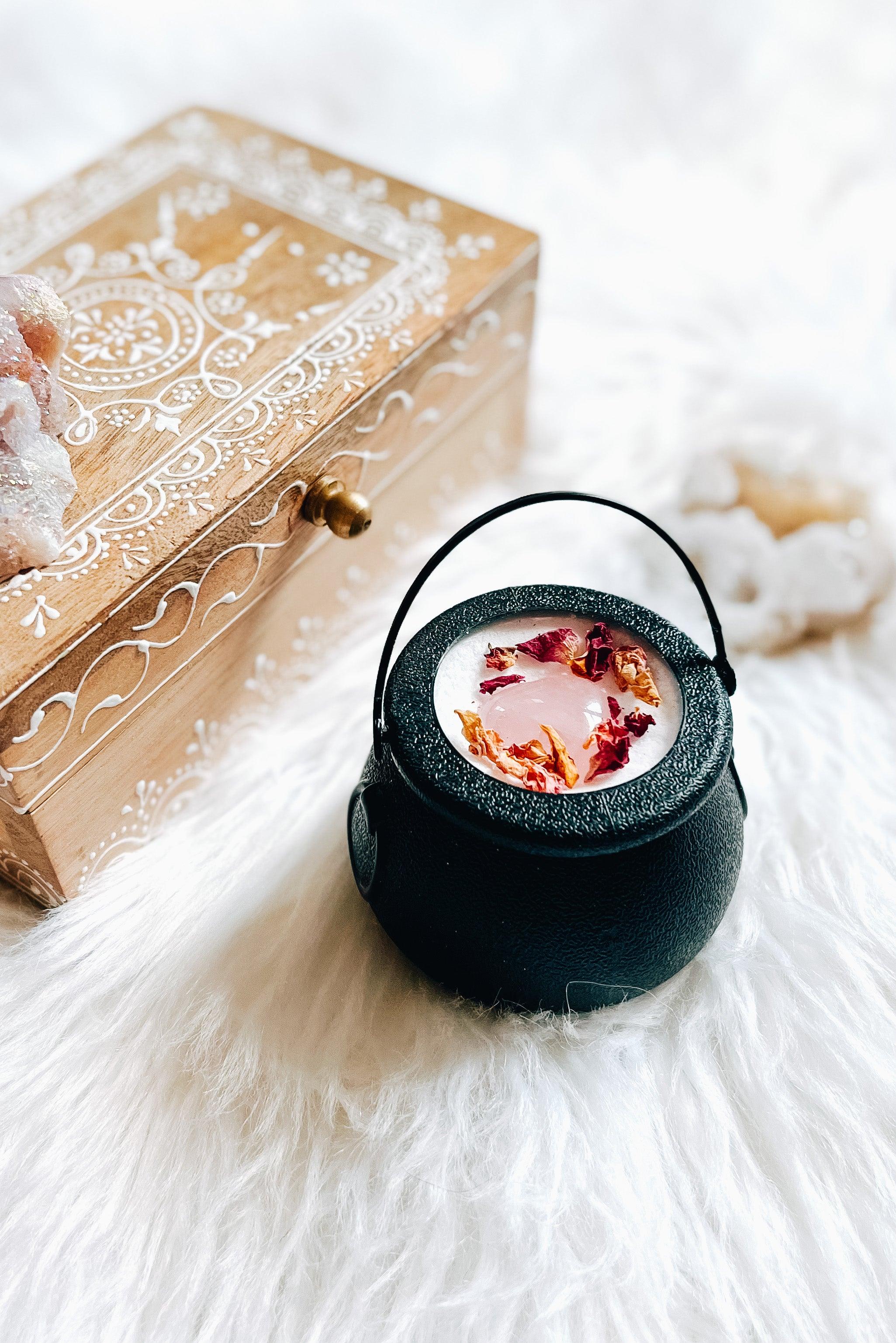 Black cauldron bath bomb with pink rose quartz heart, and dried flower petals on white fur background