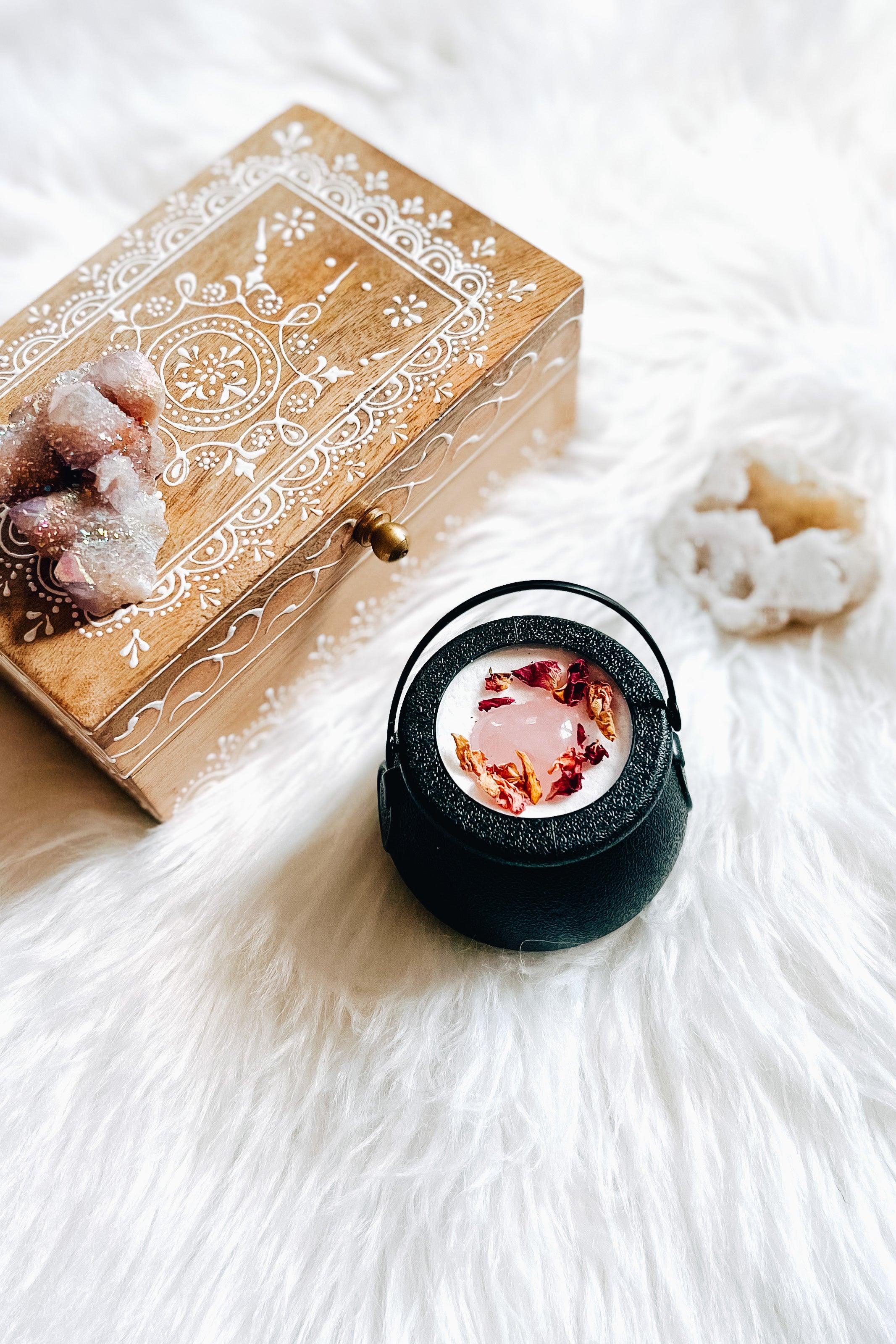 Black cauldron bath bomb with pink heart shaped crystal and dried rose petals beside ornate wooden box and white geode on white fur background