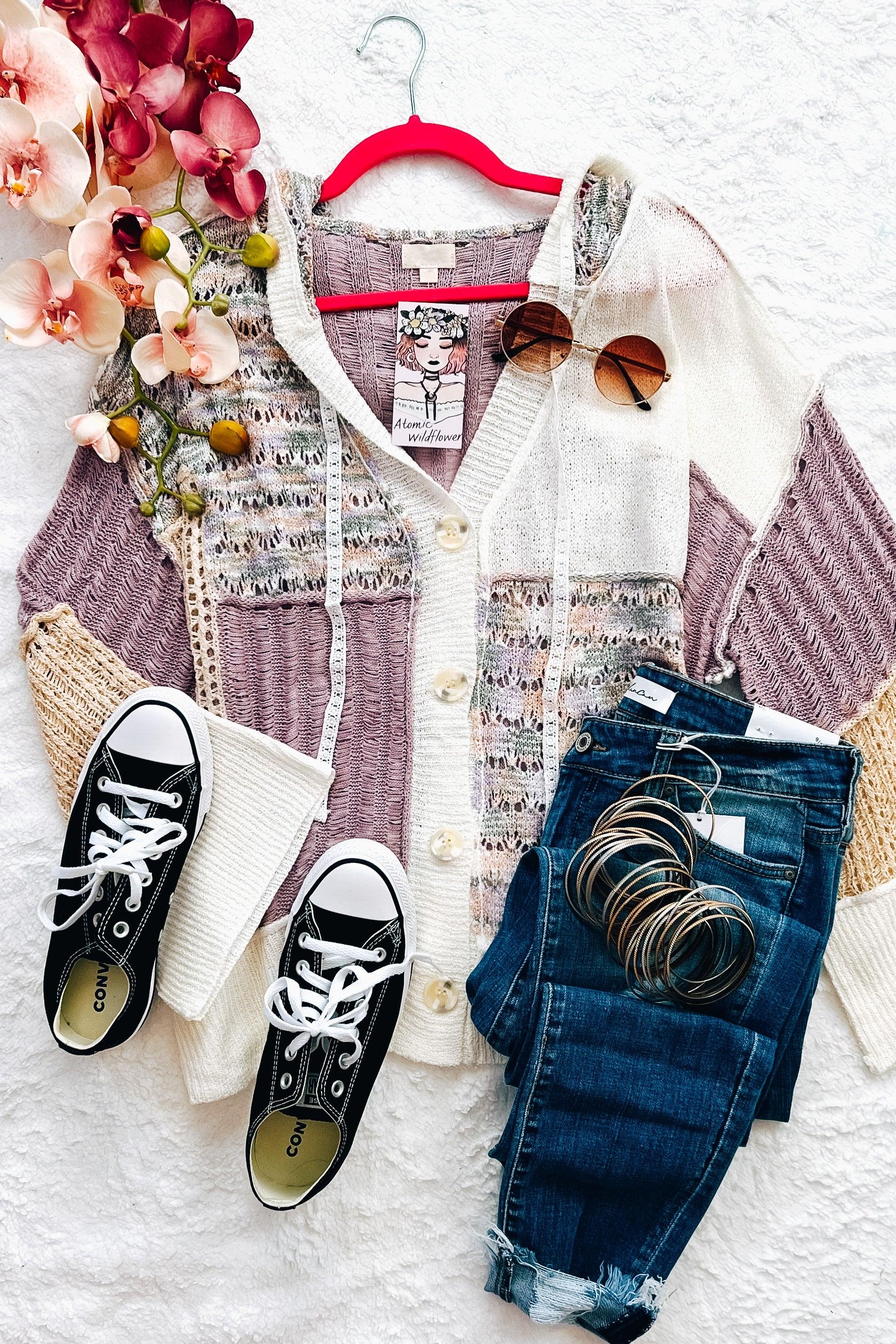Lavender cardigan with cream and beige knit panels laid out with dark jeans, black sneakers, sunglasses, and bracelets