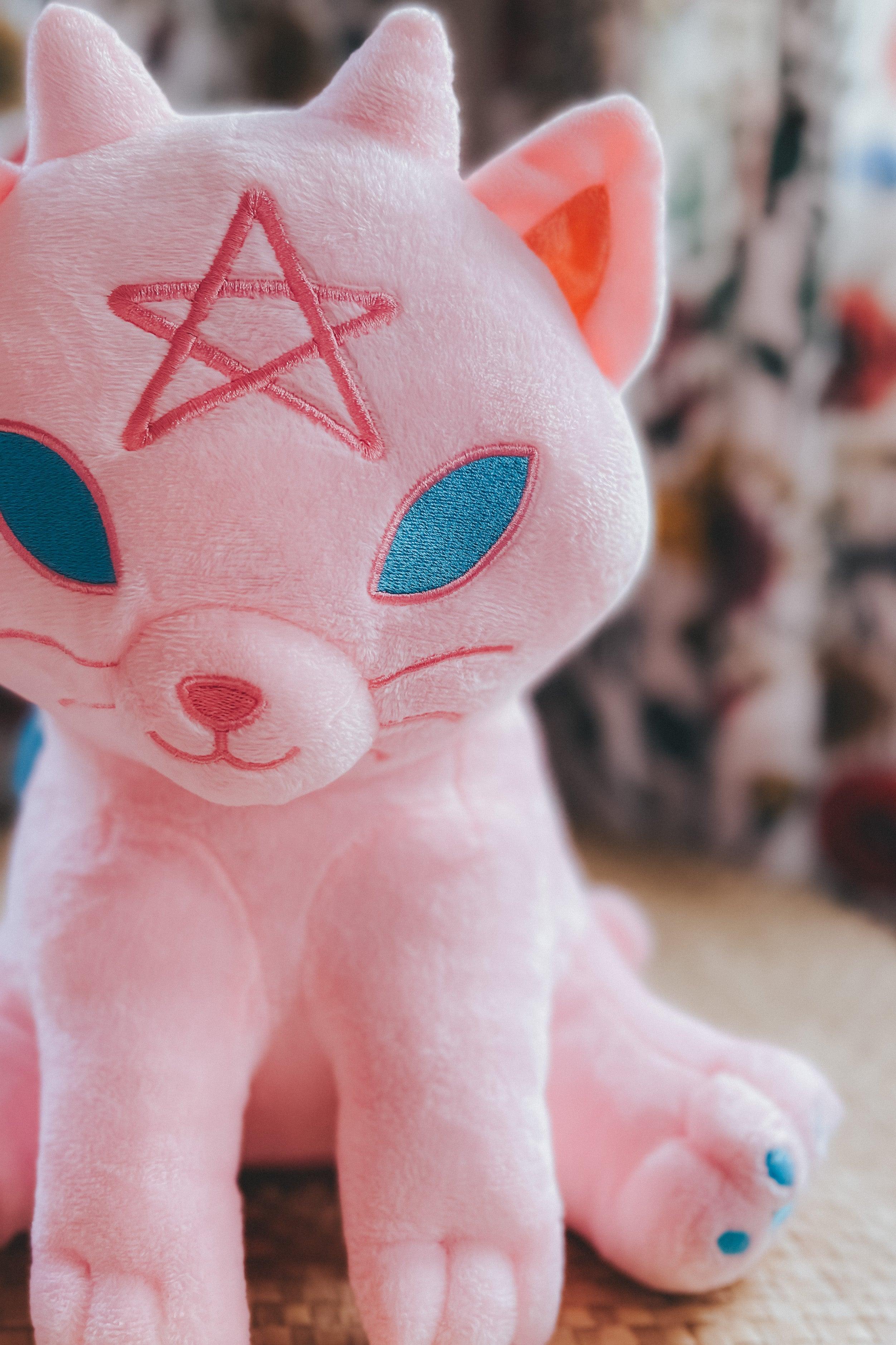 Soft cotton candy pink plush cat with stitched pentagram on forehead and teal embroidered eyes sitting on woven mat