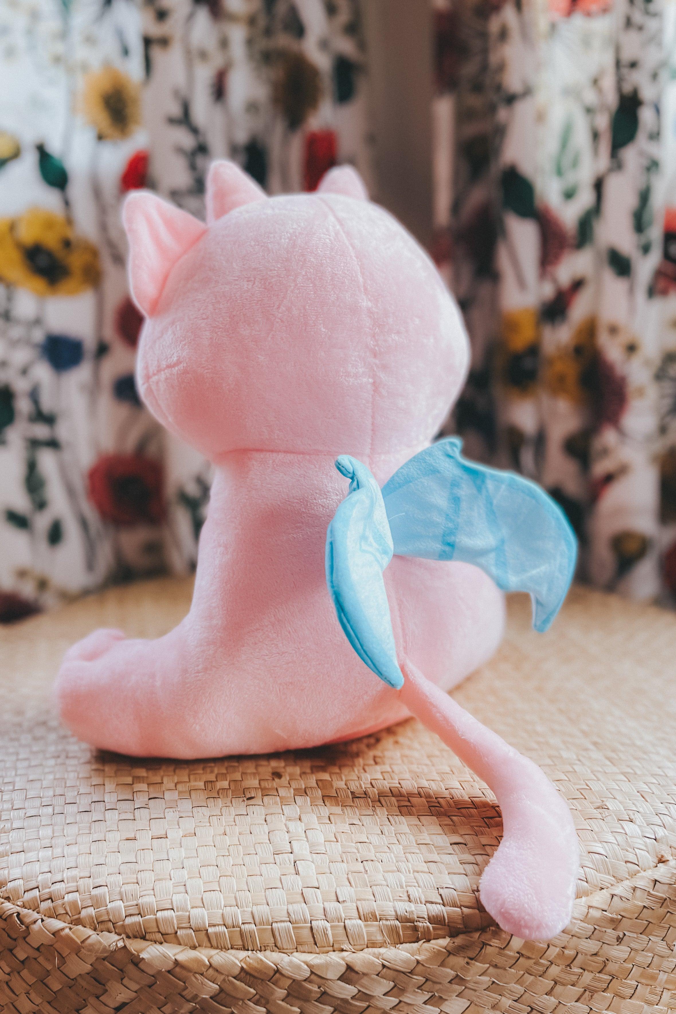 Soft cotton candy pink plush creature with blue wings and tail sitting on woven surface with floral background