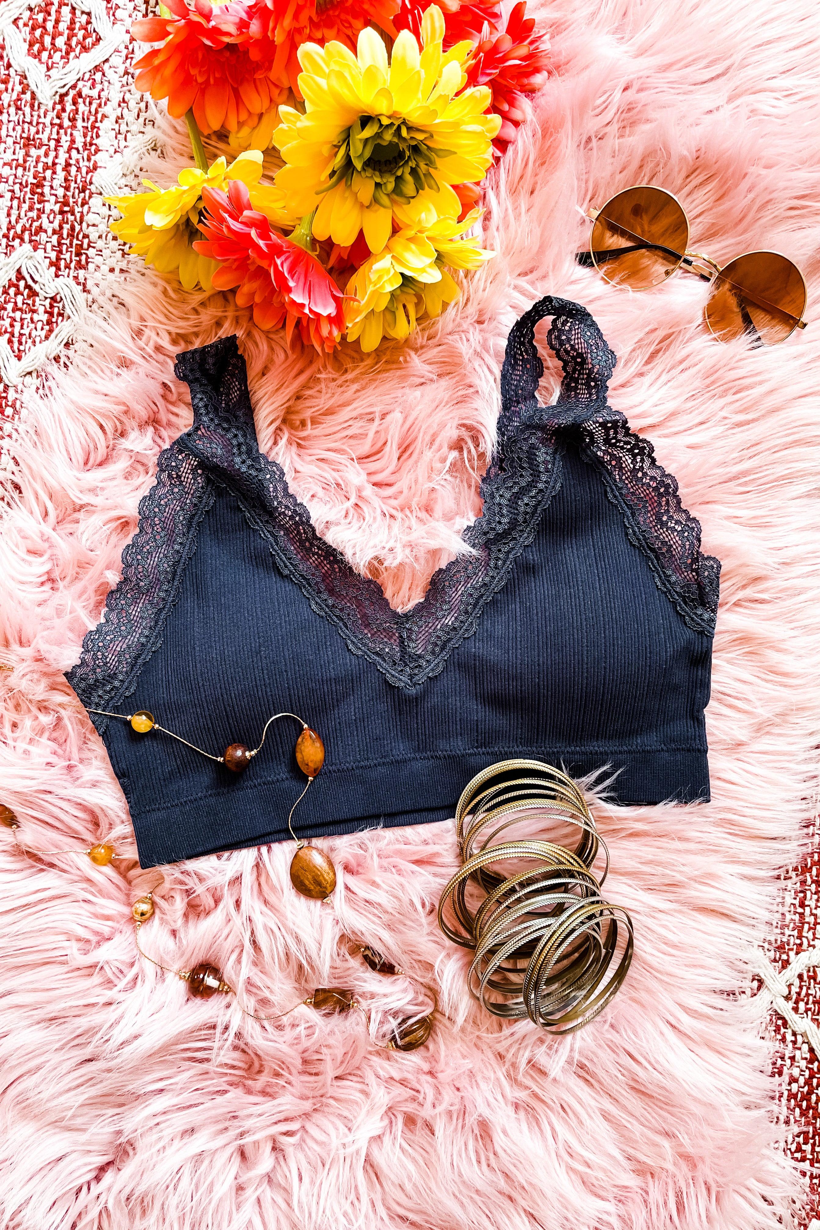 Navy lace-trimmed bralette on soft pink fur background with yellow and orange flowers, sunglasses, and jewelry accents