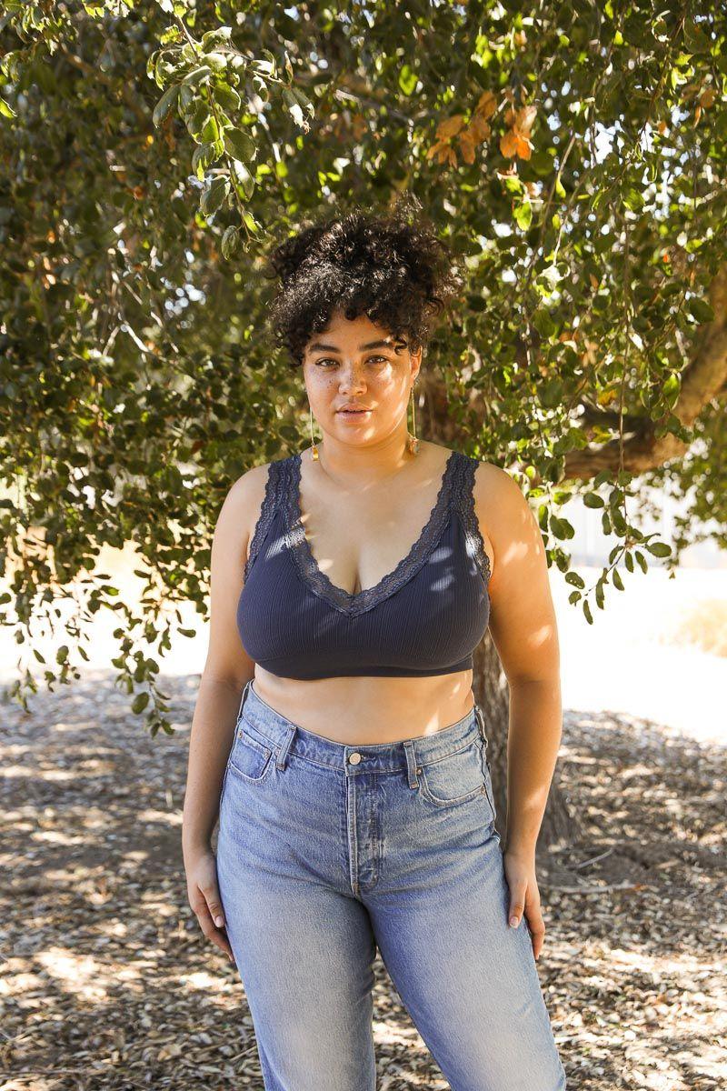 Curly-haired woman wearing navy ribbed bralette with lace trim and high-waisted light blue jeans standing outdoors by tree