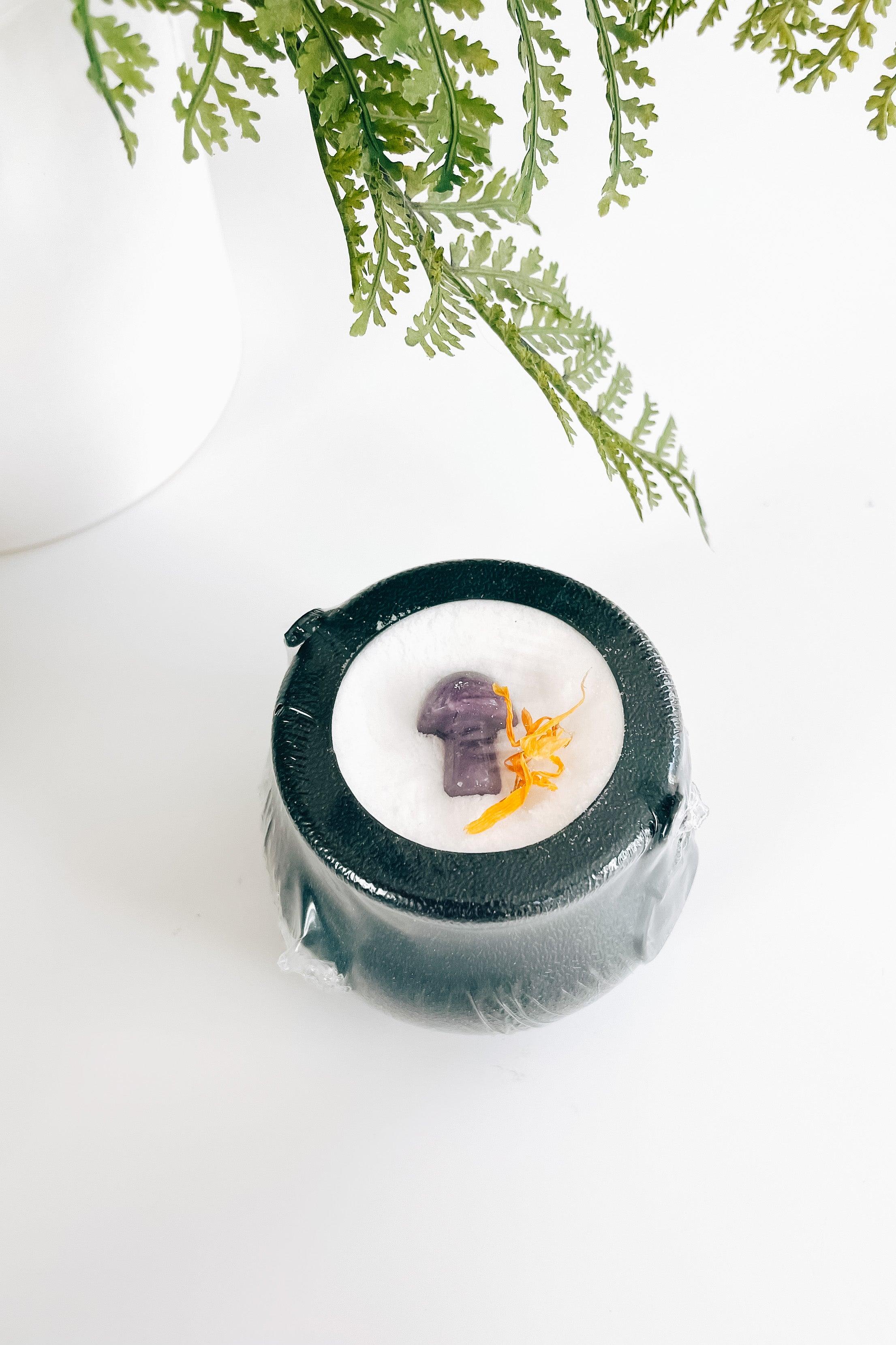 Small black cauldron bath bomb topped with a purple mushroom crystal and dried orange flower on white surface
