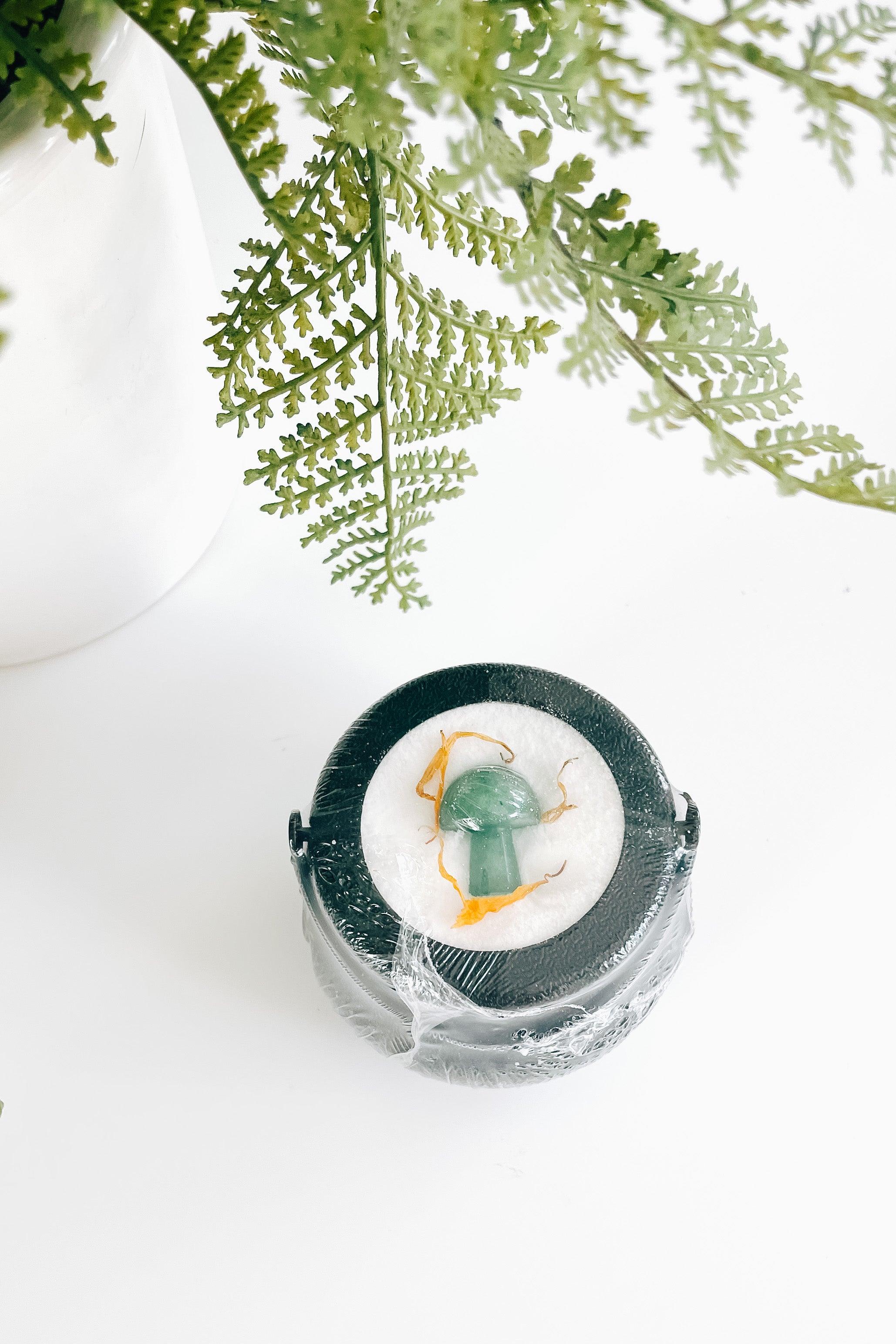 Black cauldron bath bomb with green mushroom crystal and dried orange flower under fern leaves on white surface
