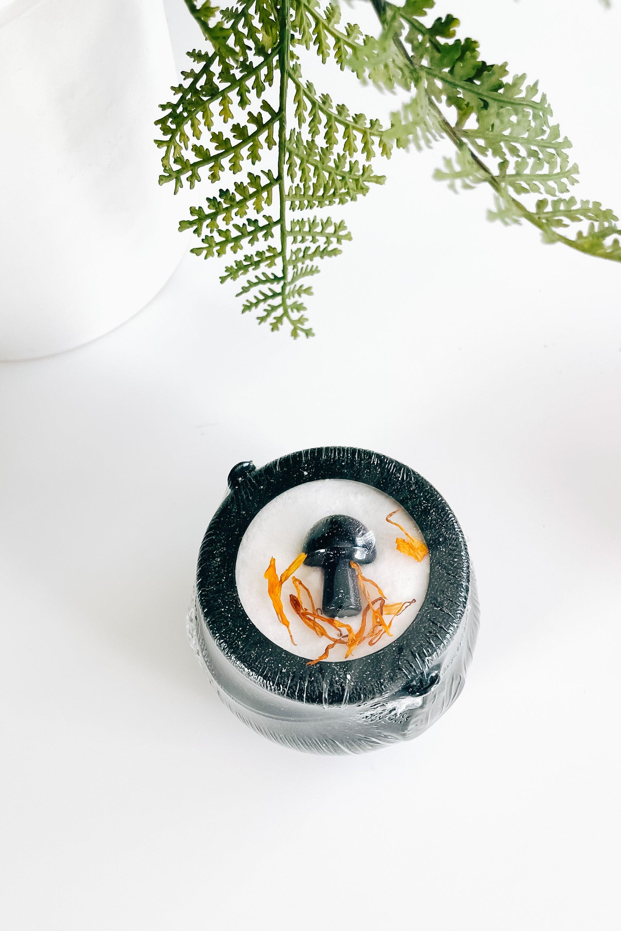 Black cauldron bath bomb with small black mushroom crystal and orange dried petals under green fern leaves