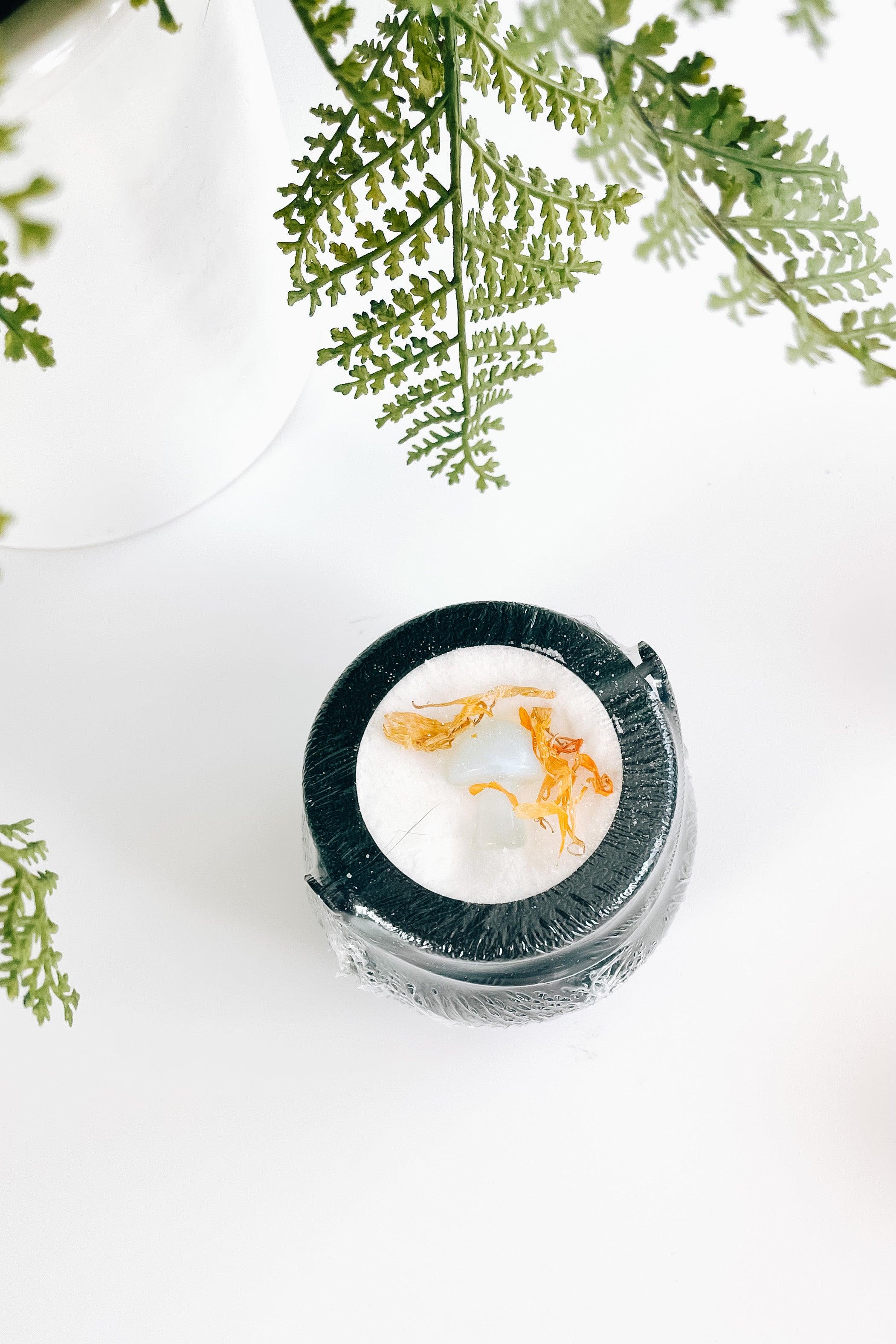 Small black cauldron bath bomb holding a white mushroom crystal topped with dried golden flower petals under green fern leaves