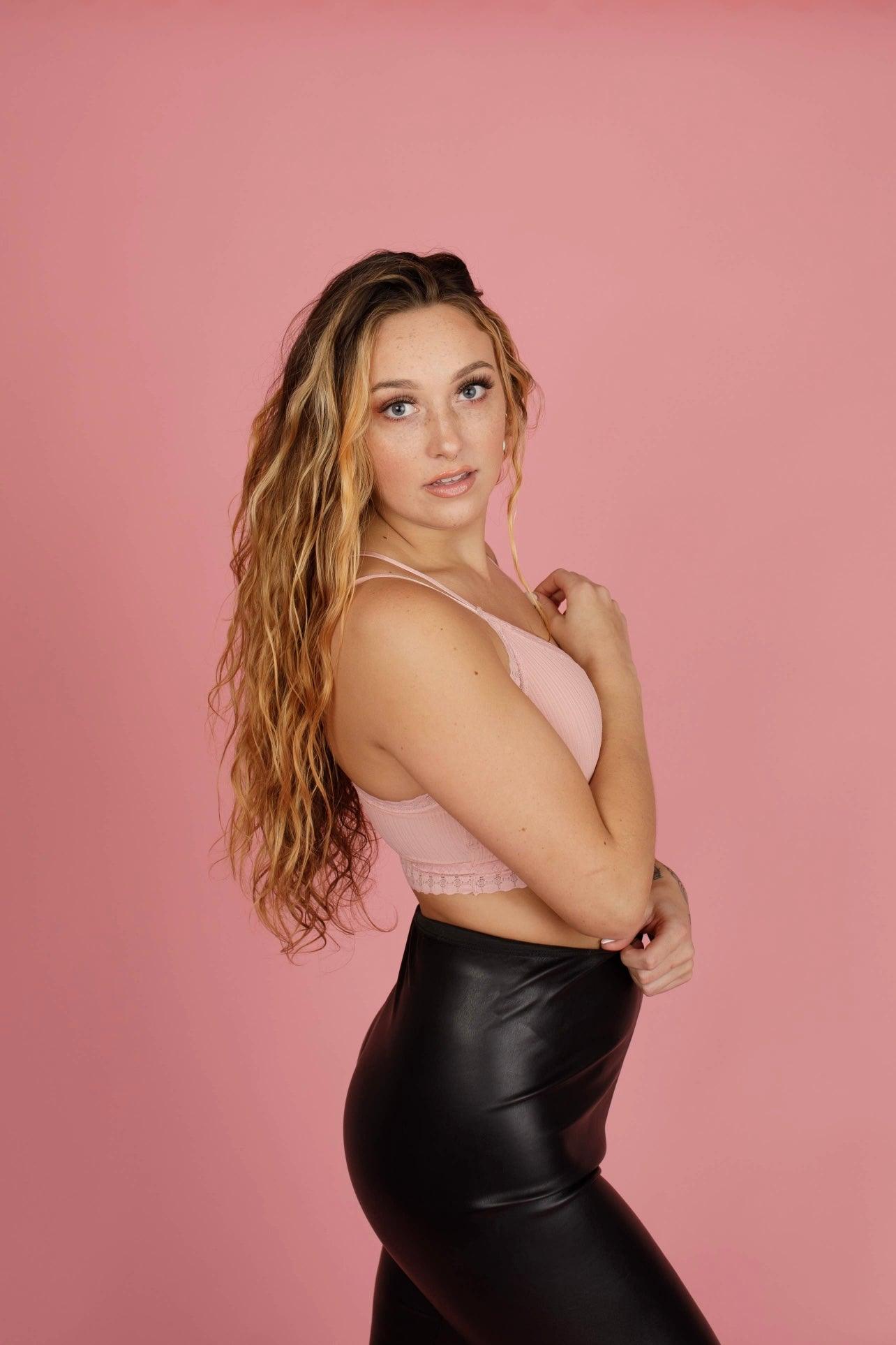 Woman with long wavy hair wearing a soft pink lace bralette and high-waisted black faux leather pants against pink background