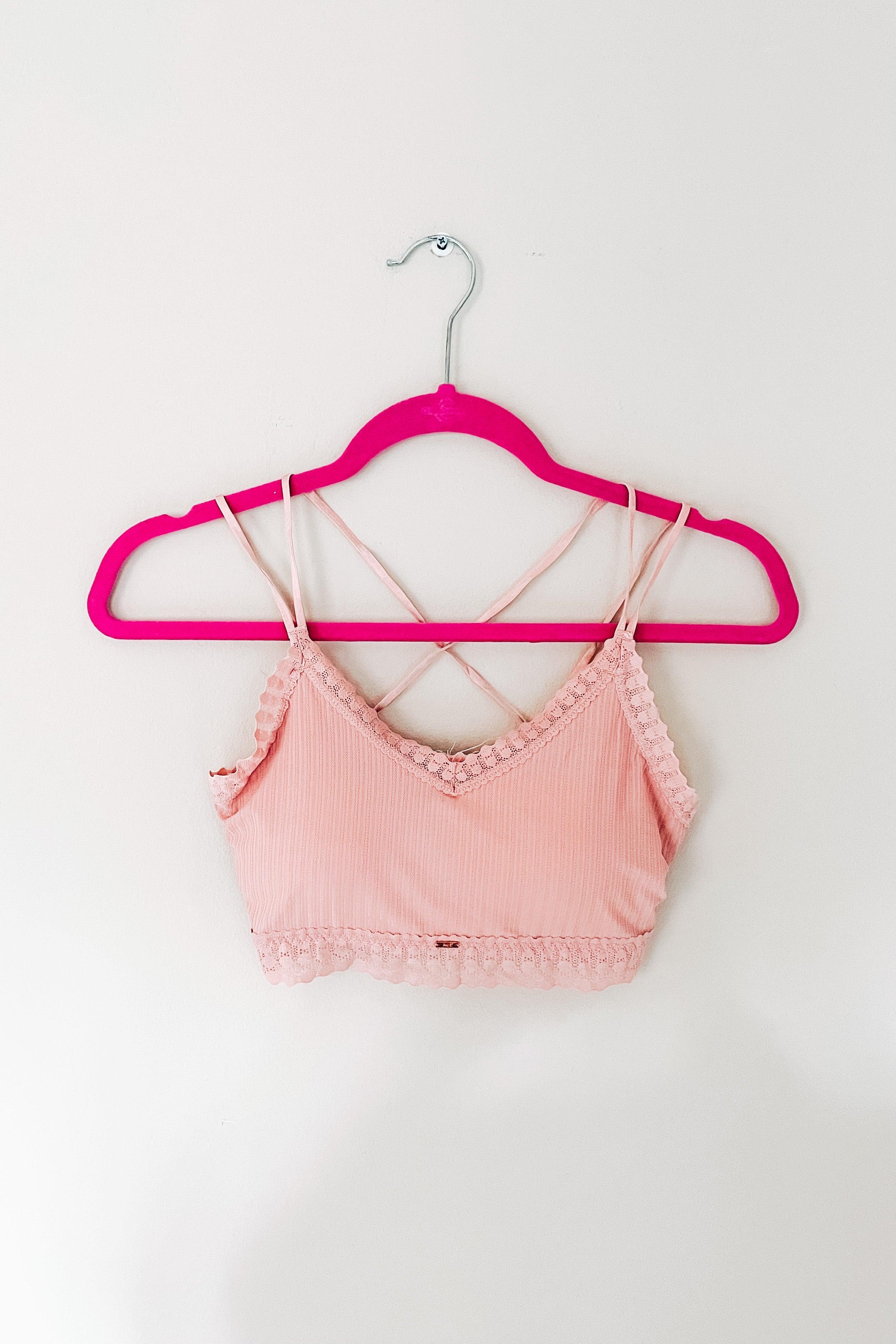 Light pink ribbed bralette with delicate lace trim and thin crisscross straps, displayed on a pink hanger against white background