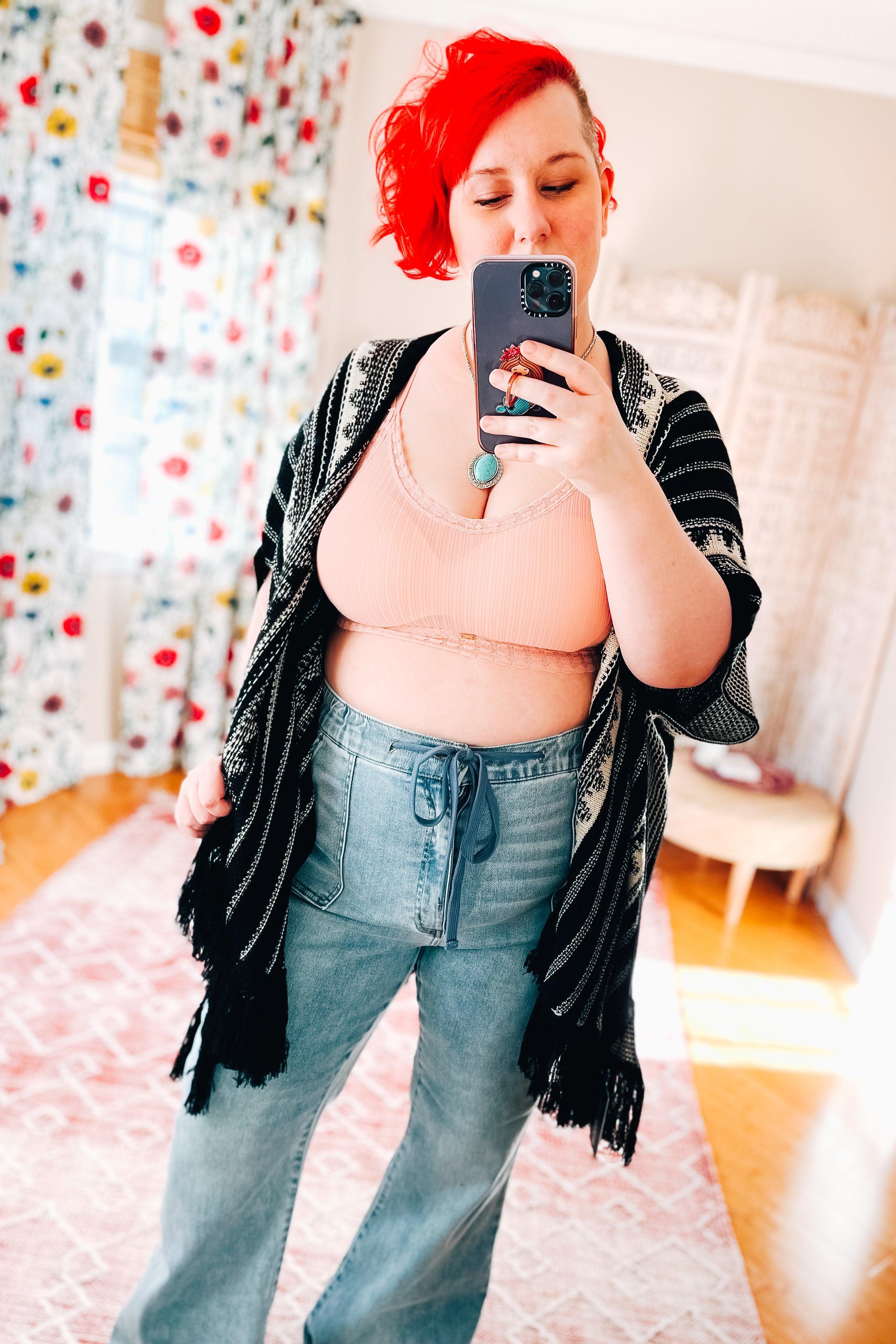 Woman with bright red hair wearing pink lace-trimmed bralette and high-waisted loose jeans taking a mirror selfie