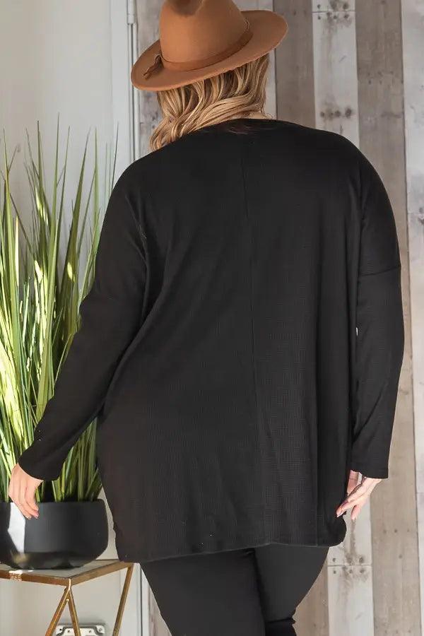 Back view of a person wearing a loose black oversized tunic with subtle seam detail and long sleeves indoors