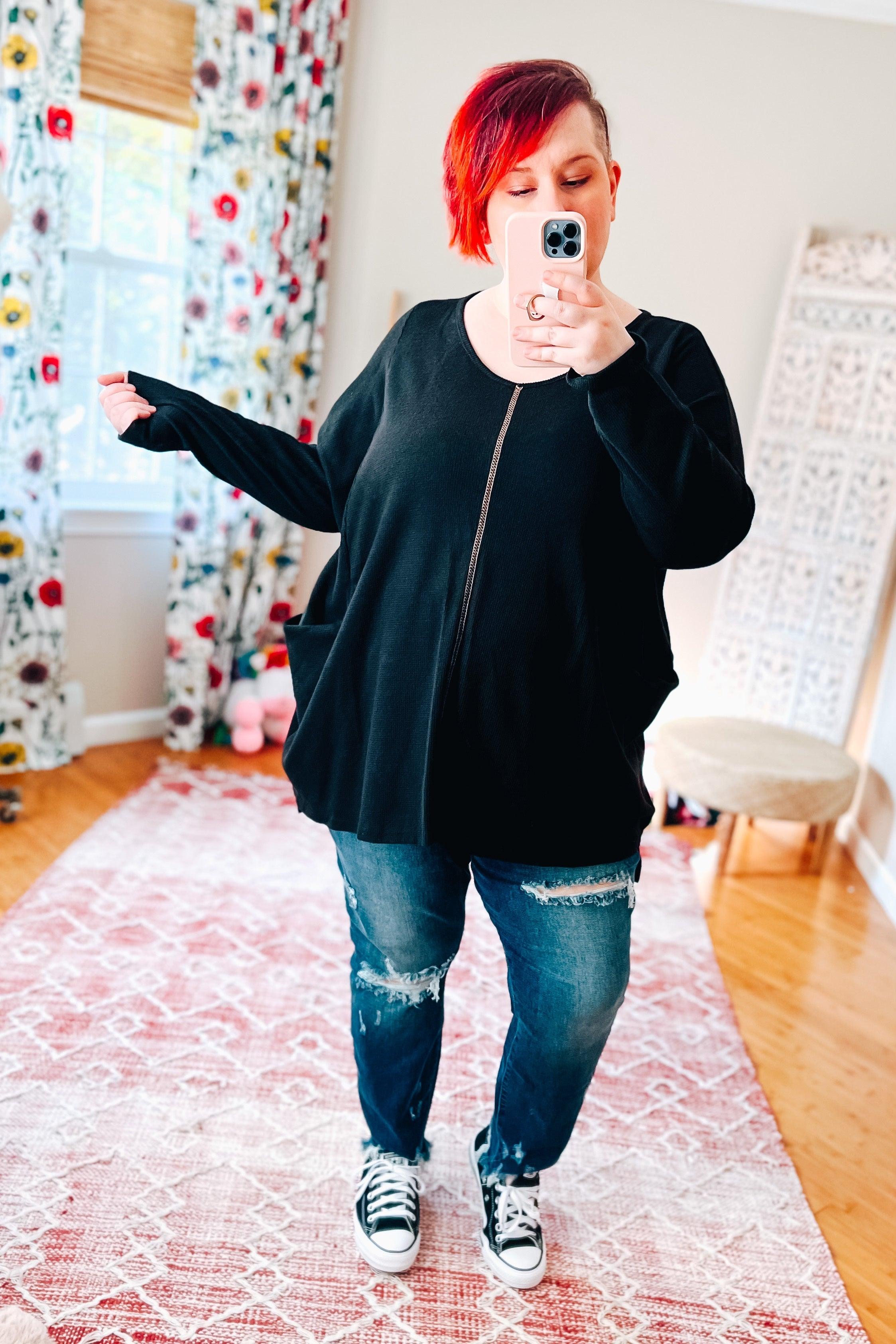 Woman taking a mirror selfie wearing a loose black thermal tunic with pockets and distressed blue jeans indoors