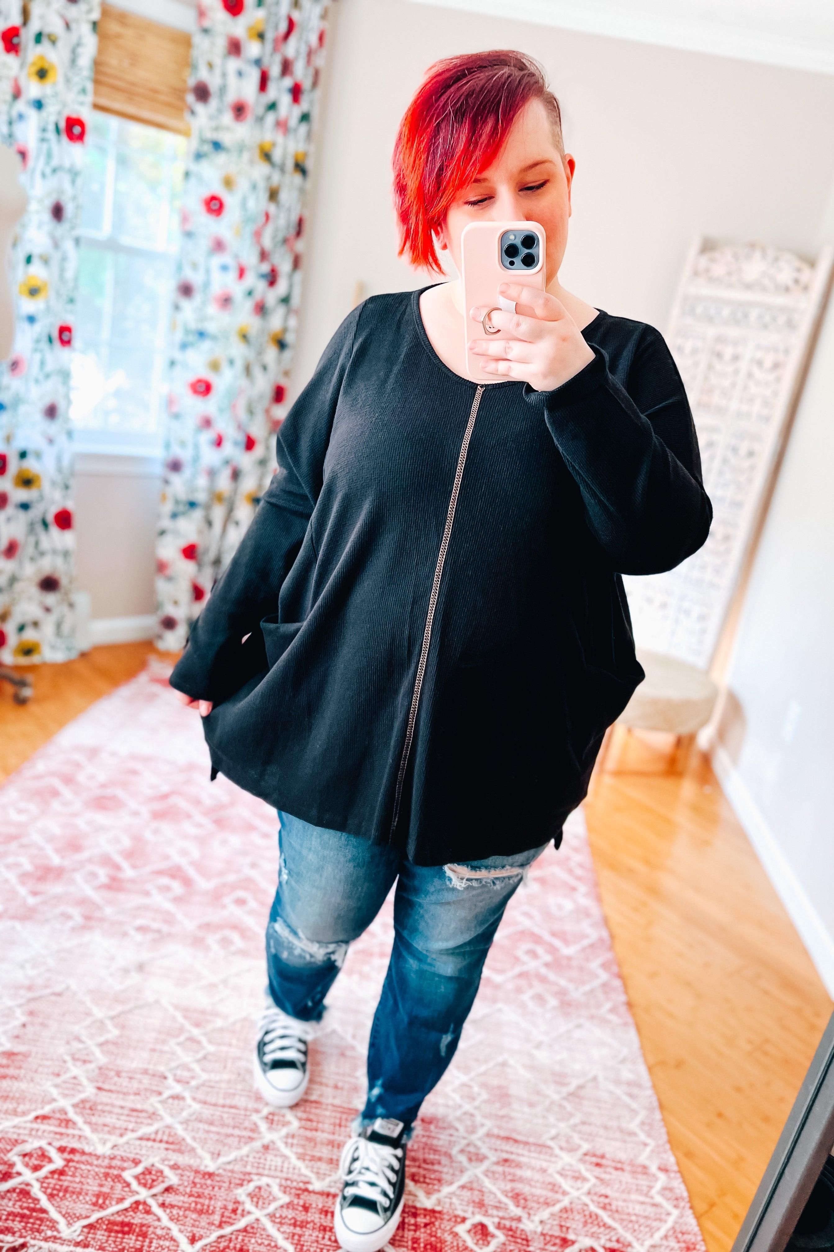 Woman taking a mirror selfie wearing a loose black long-sleeve tunic with front pockets and ripped blue jeans indoors