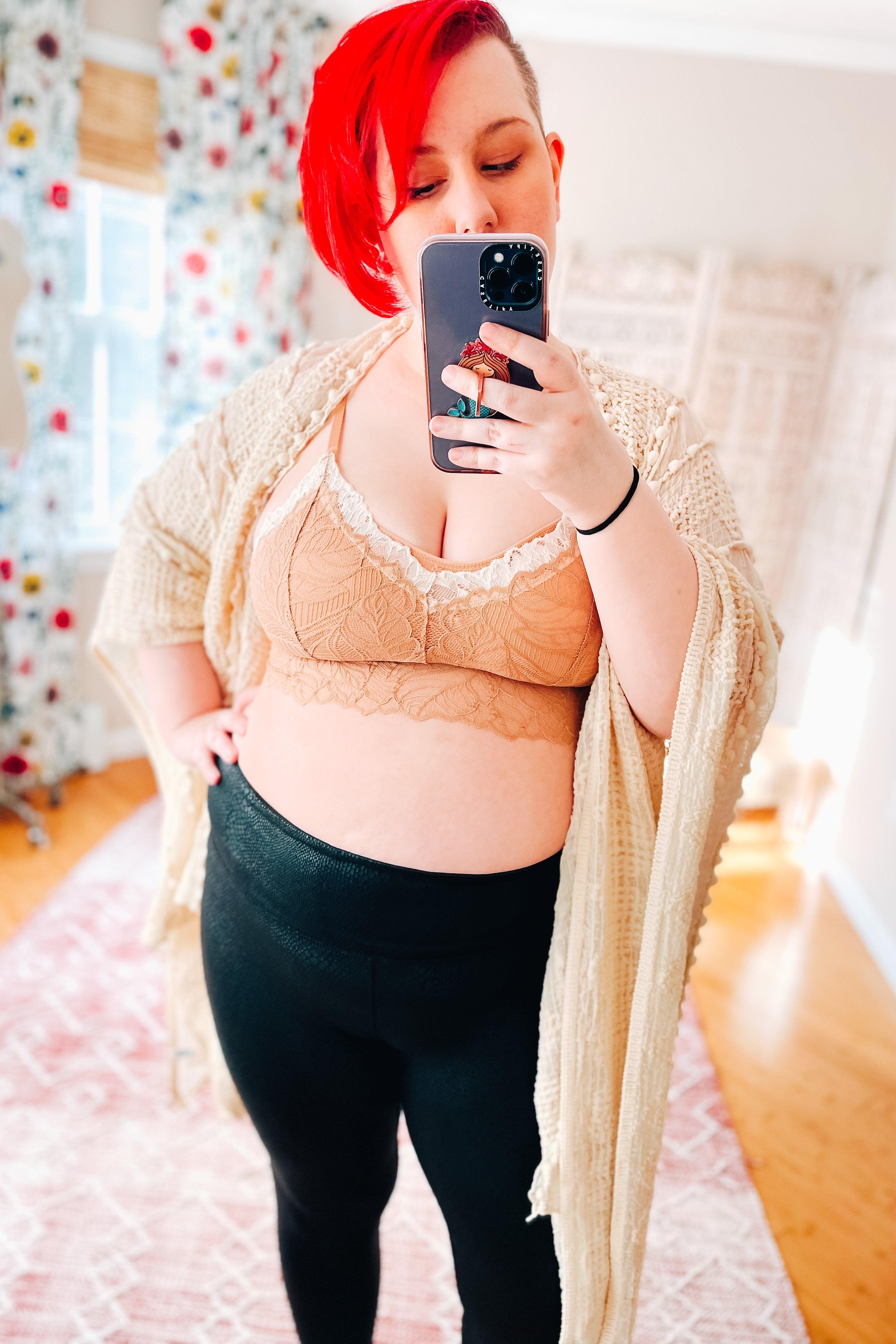 Woman with bright red hair wearing a brick-colored lace bralette with floral details, black leggings, and a cream knitted cardigan taking a mirror selfie in a cozy room.