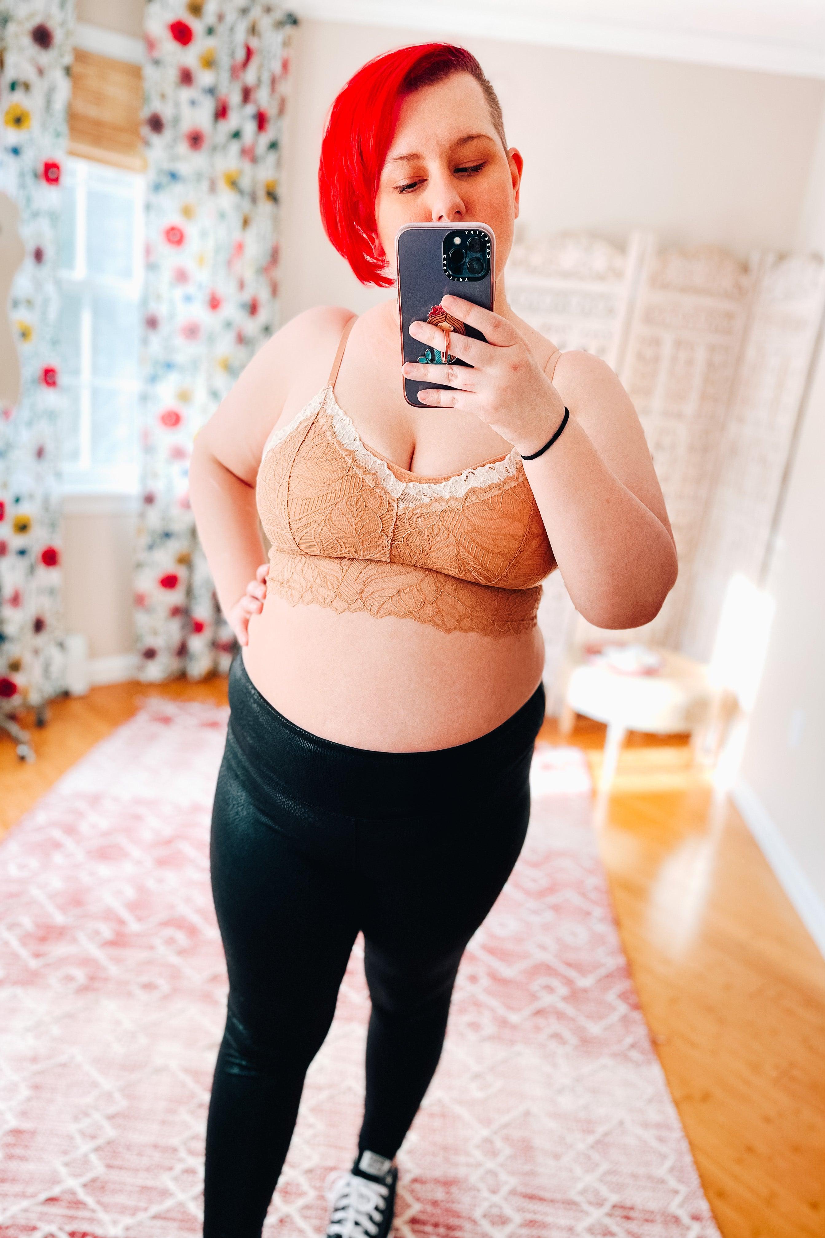 Woman with bright red hair wearing a brick-colored lace bralette with delicate cream trim and black leggings taking a mirror selfie in a sunlit room.
