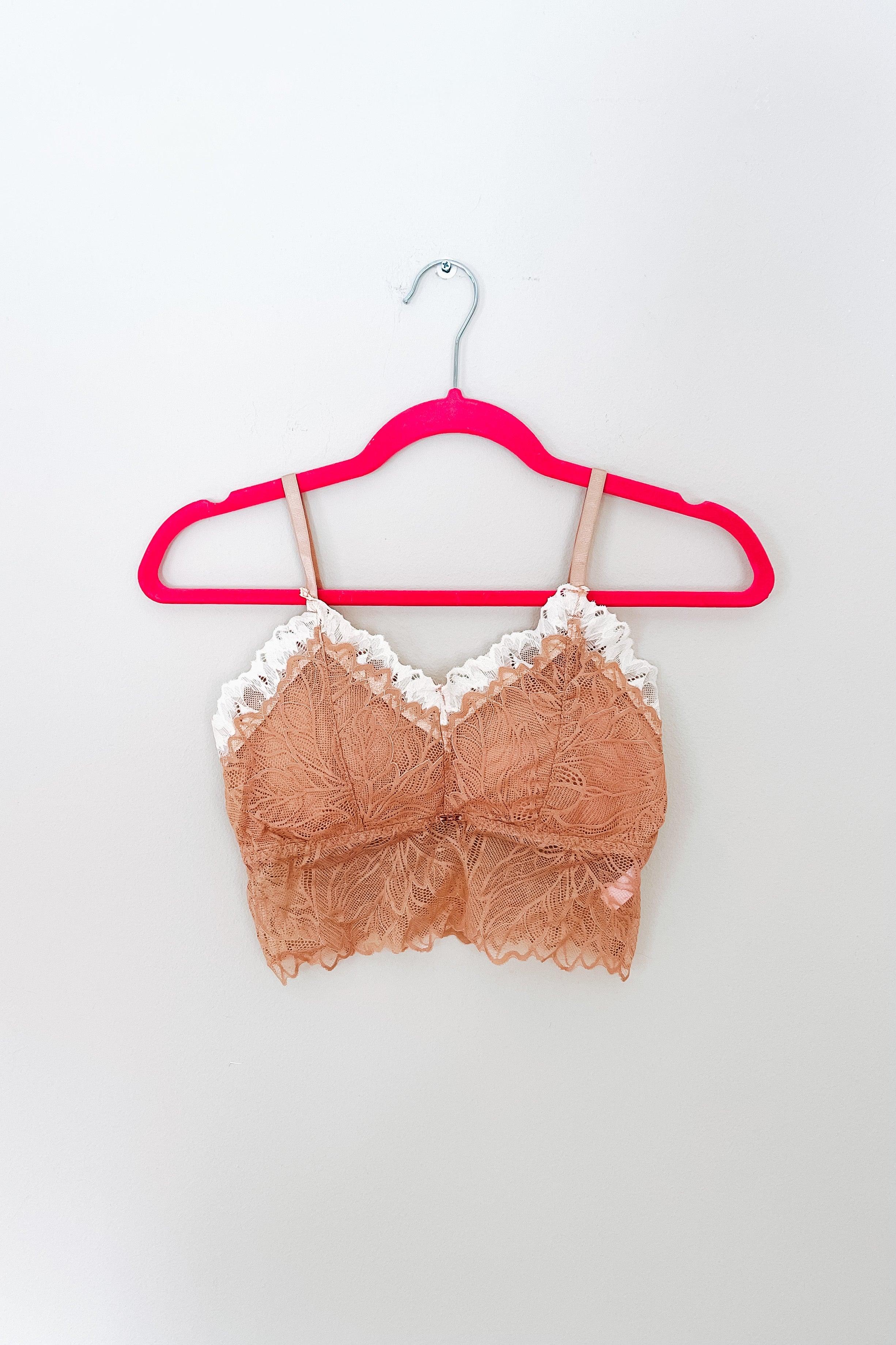 Delicate brick-colored lace bralette with white scalloped trim, hanging on a bright pink hanger against a white wall background.