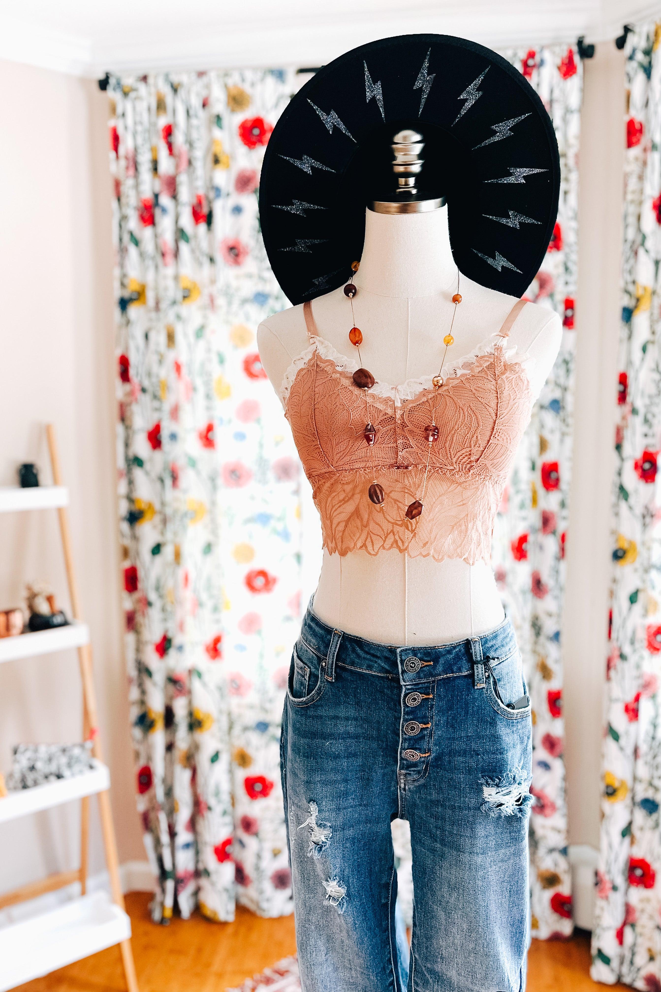 Brick-colored lace bralette with scalloped edges displayed on a mannequin styled with a layered amber bead necklace and distressed blue jeans against floral curtains.