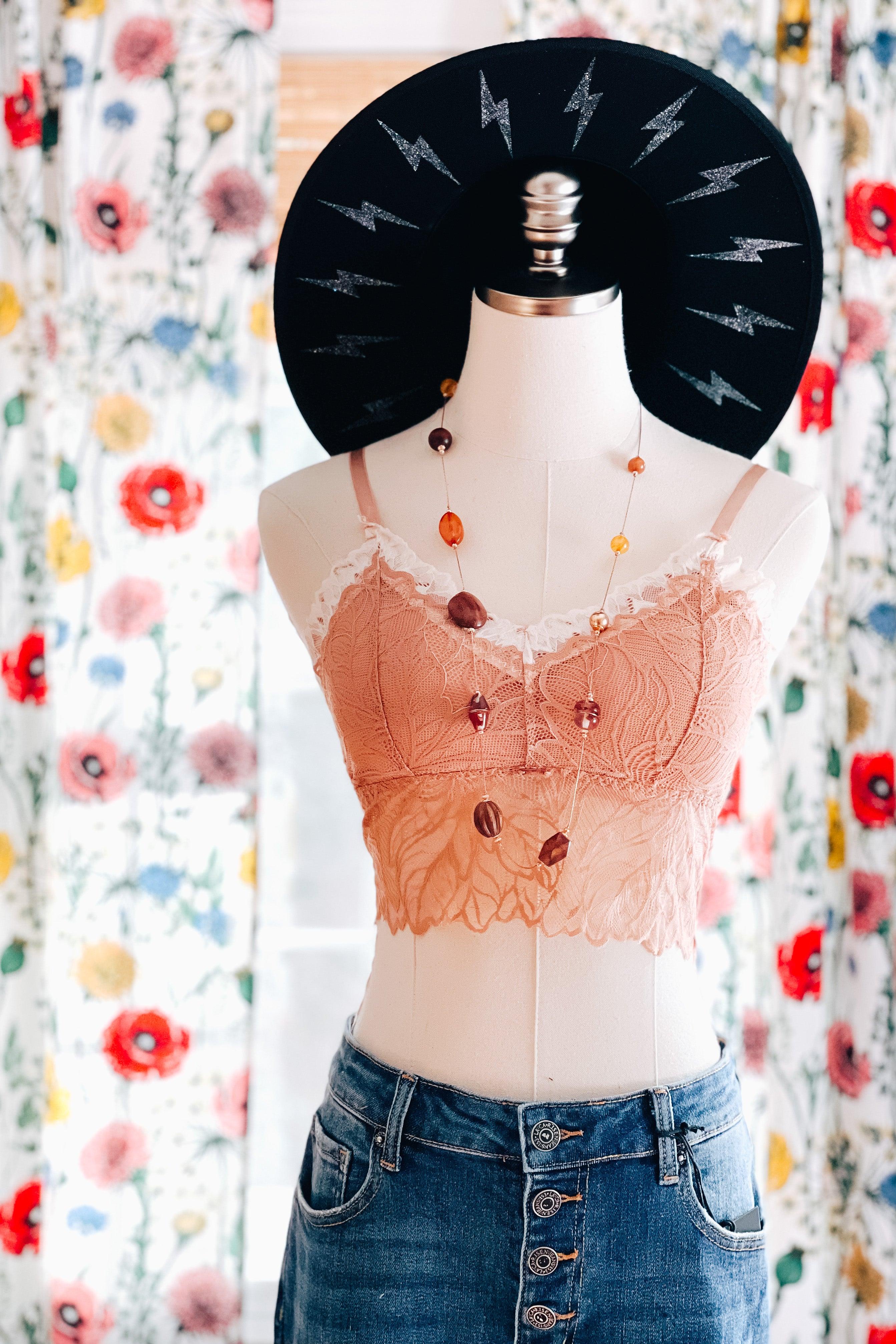 Brick-colored lace bralette with white scalloped trim styled on a mannequin wearing layered amber bead necklace, high-waisted blue denim jeans, and a black hat with silver lightning bolts against floral curtains.
