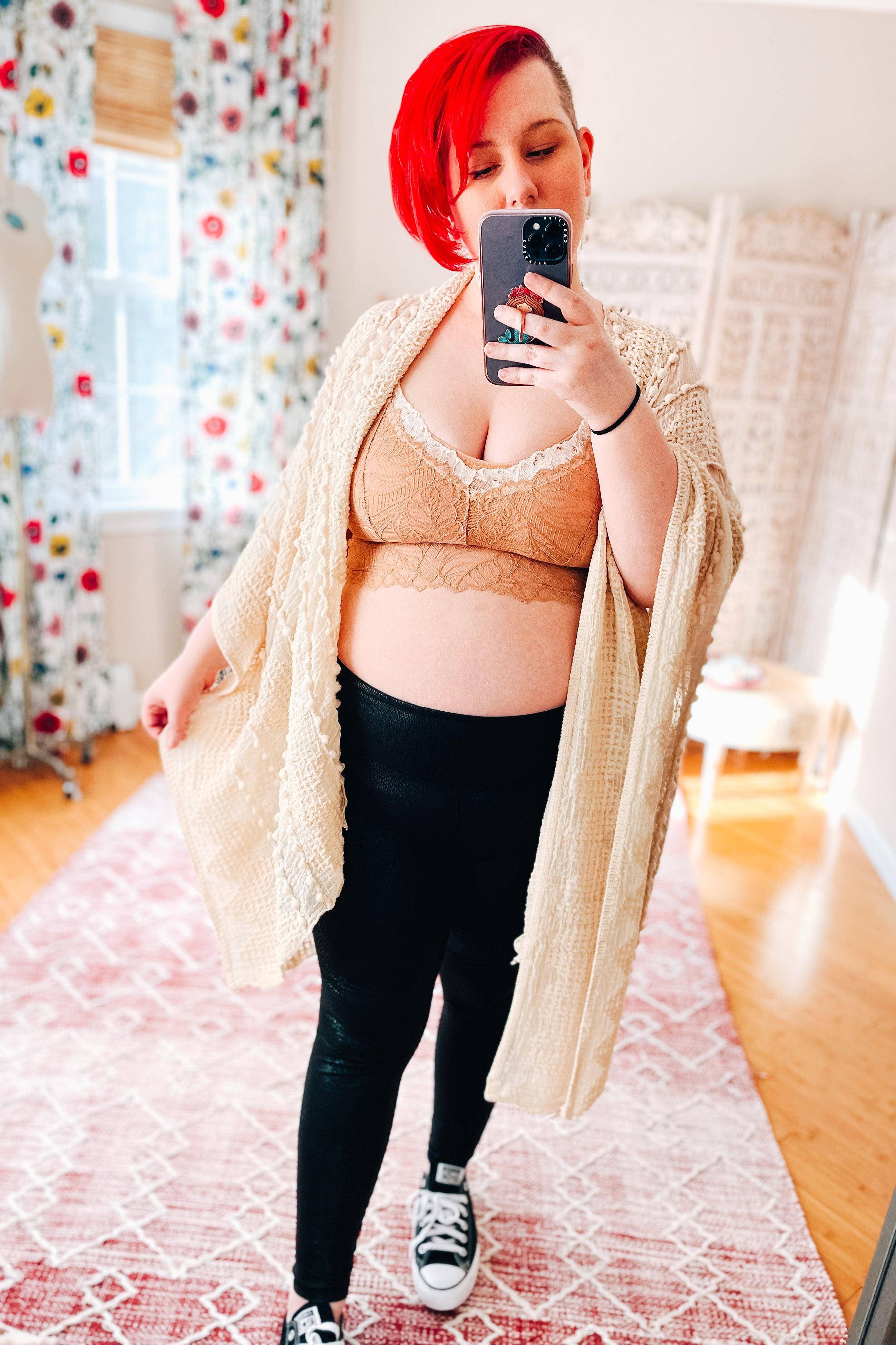 Woman with bright red hair wearing a brick-colored lace bralette with white trim, black leggings, and a cream knitted cardigan, taking a mirror selfie in a bright room.
