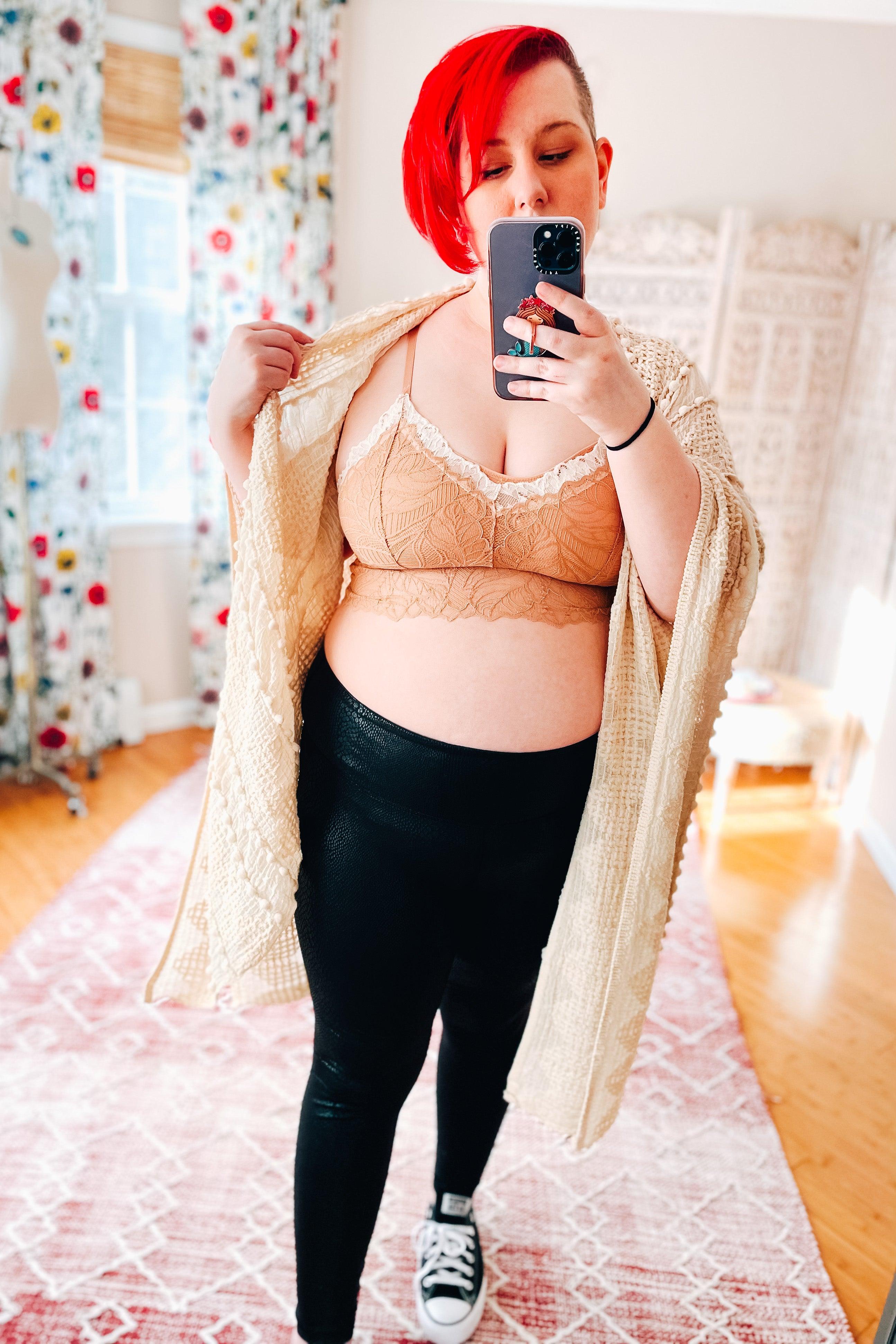 Woman with bright red hair wearing a brick-colored lace bralette with cream scalloped trim, black leggings, cream knitted cardigan, and black sneakers taking a mirror selfie in a bright room.