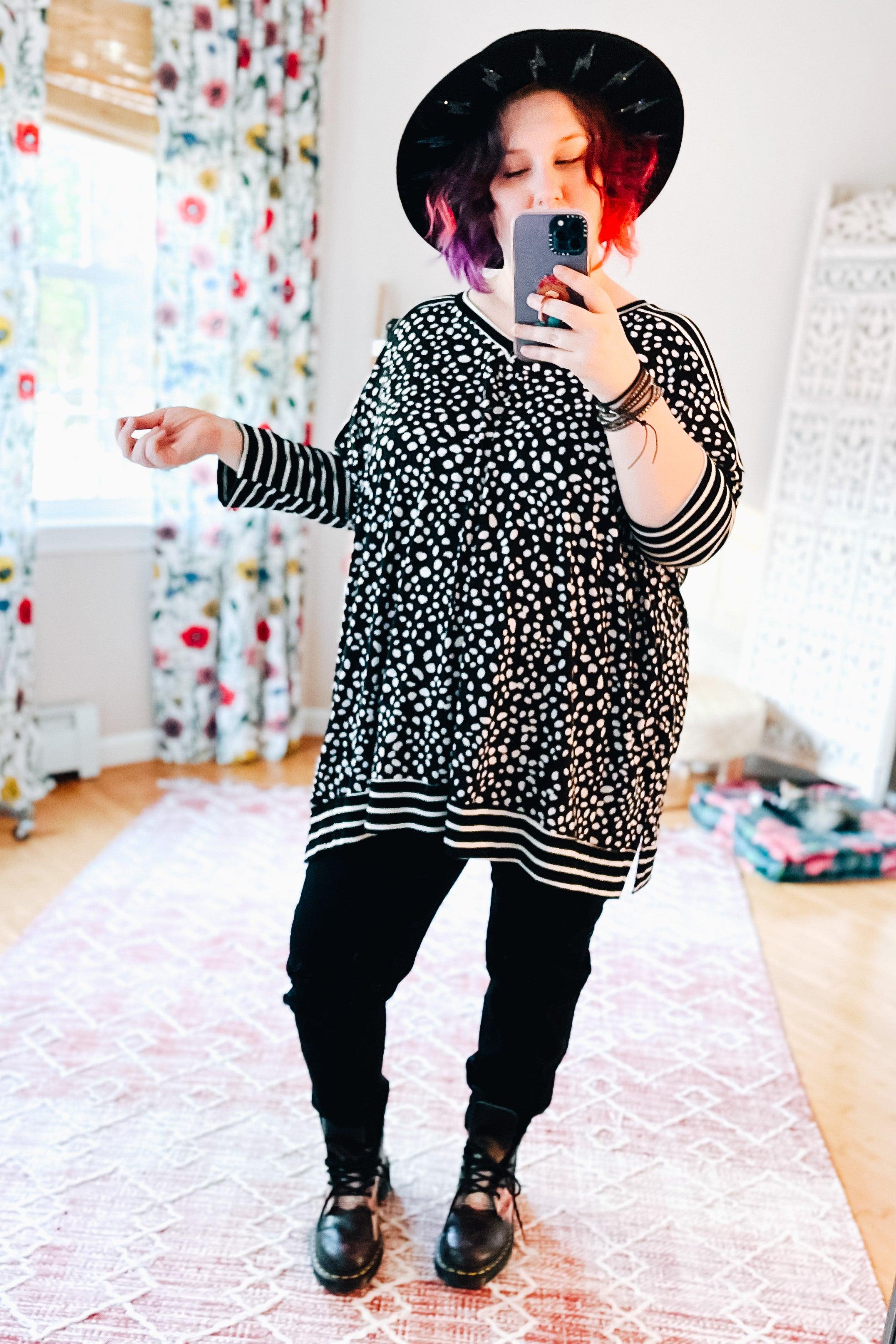 Woman wearing a loose black tunic with white irregular dots and striped sleeves and hem, paired with black pants, boots, and a black hat, taking a mirror selfie in a sunlit room.
