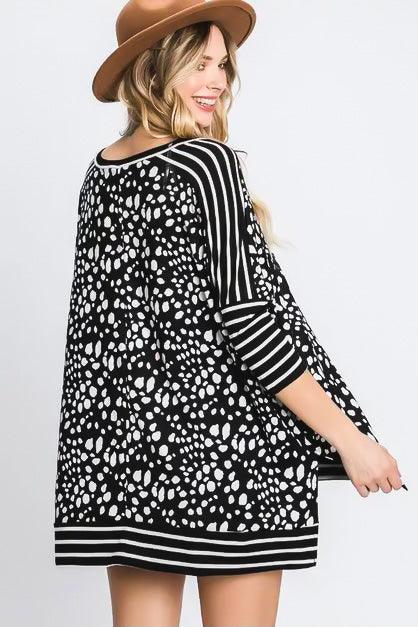 Woman wearing a flowy black tunic featuring white abstract spots and contrasting black and white striped sleeves and hem, smiling and looking back.