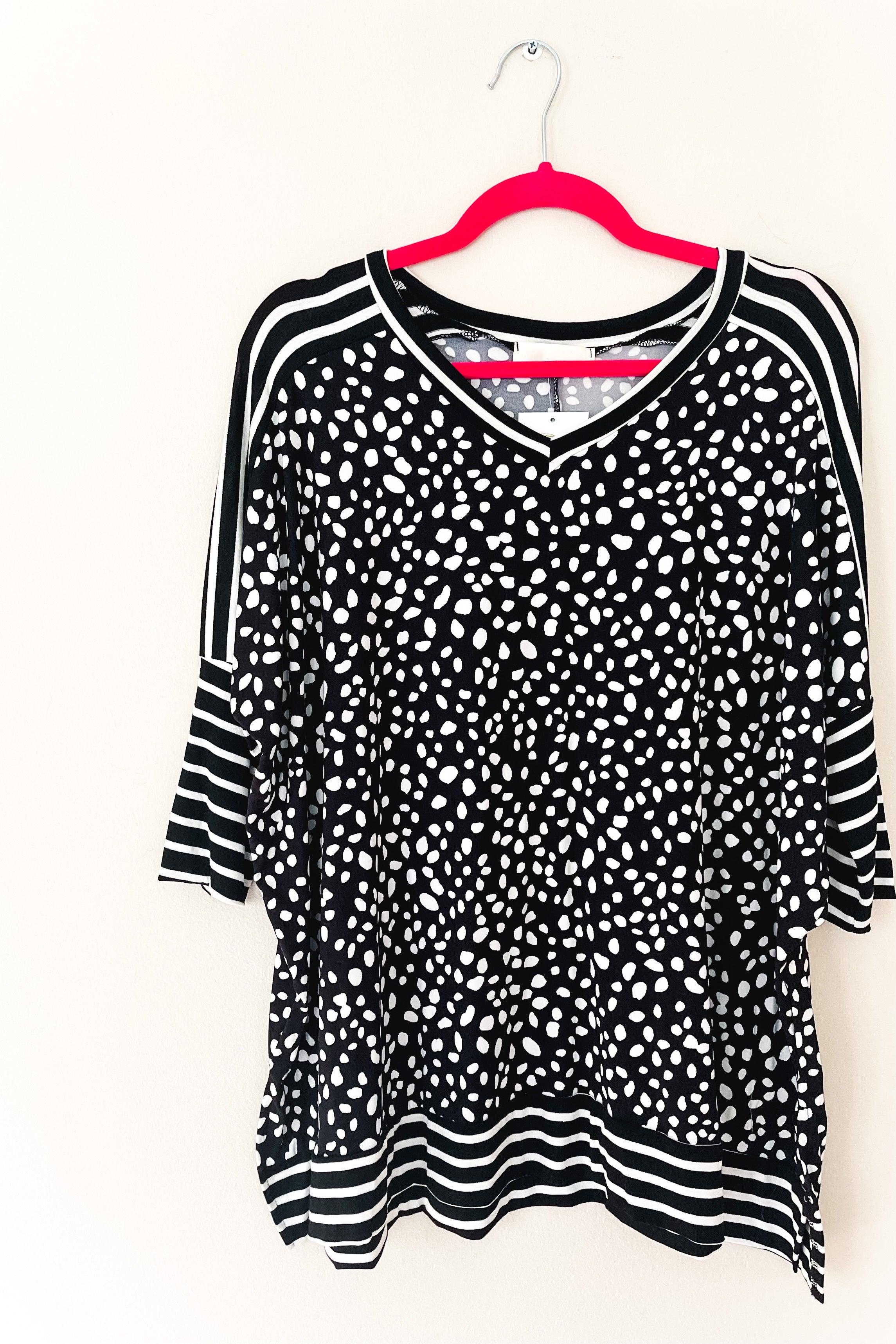 Black tunic with white irregular spots, contrasting striped mid sleeves, hem, and neckline, hanging on a pink hanger against a white background.