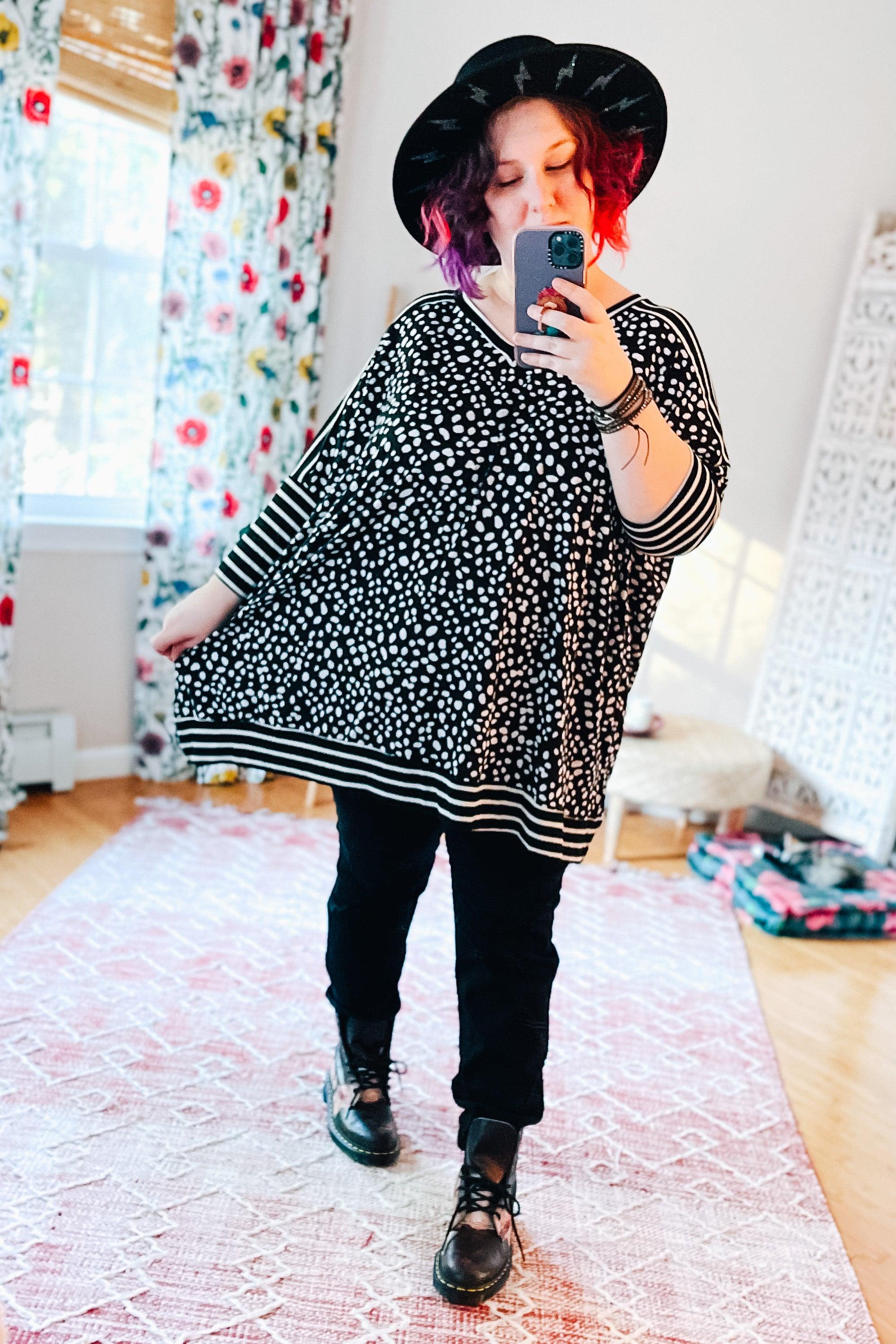 Woman taking a mirror selfie wearing a black patterned tunic with striped mid sleeves, black pants, black boots, and a black wide-brim hat in a bright room.