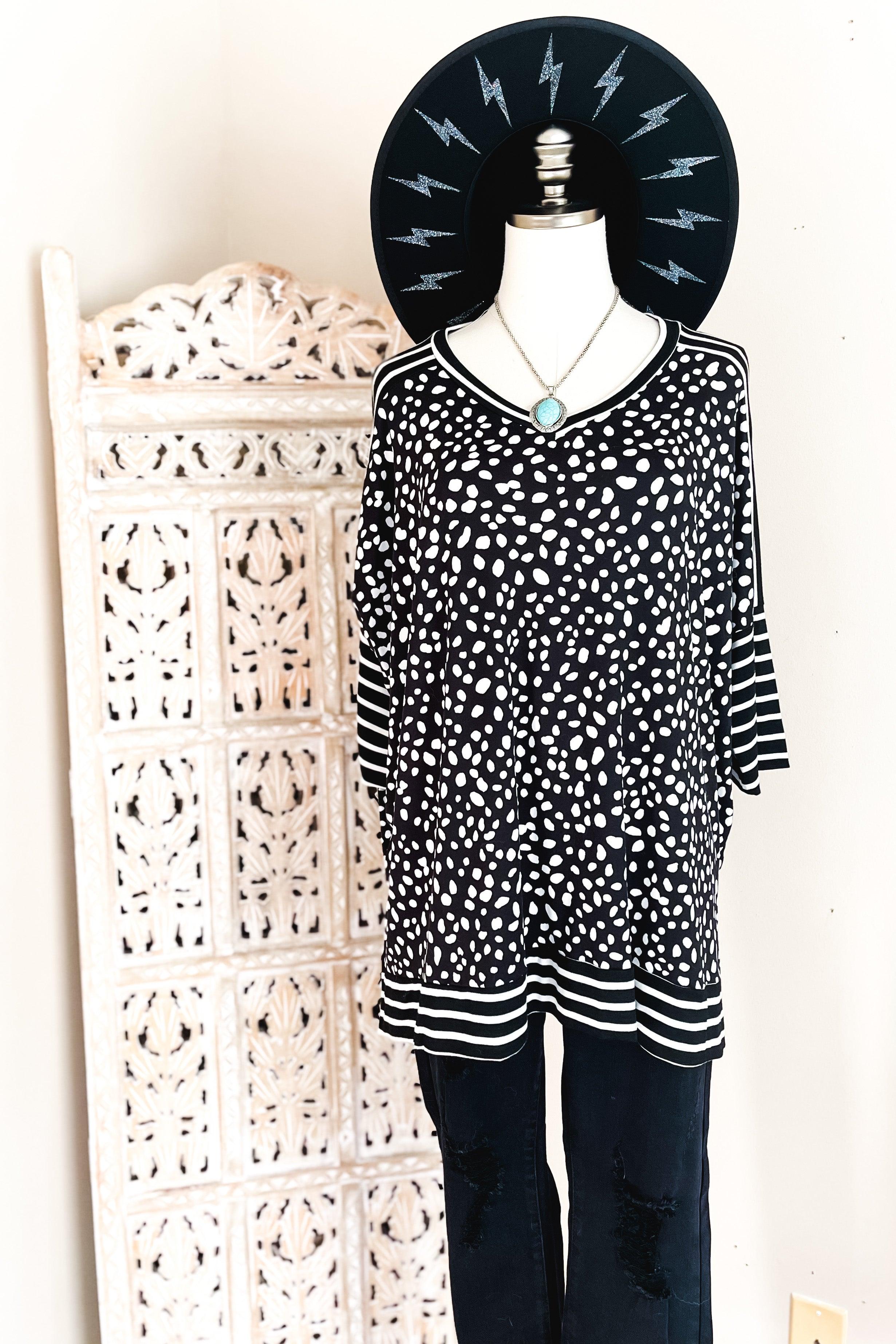 Loose black tunic with irregular white spots and contrasting striped mid sleeves, hem, and neckline styled with a turquoise pendant necklace and wide-brim black hat.