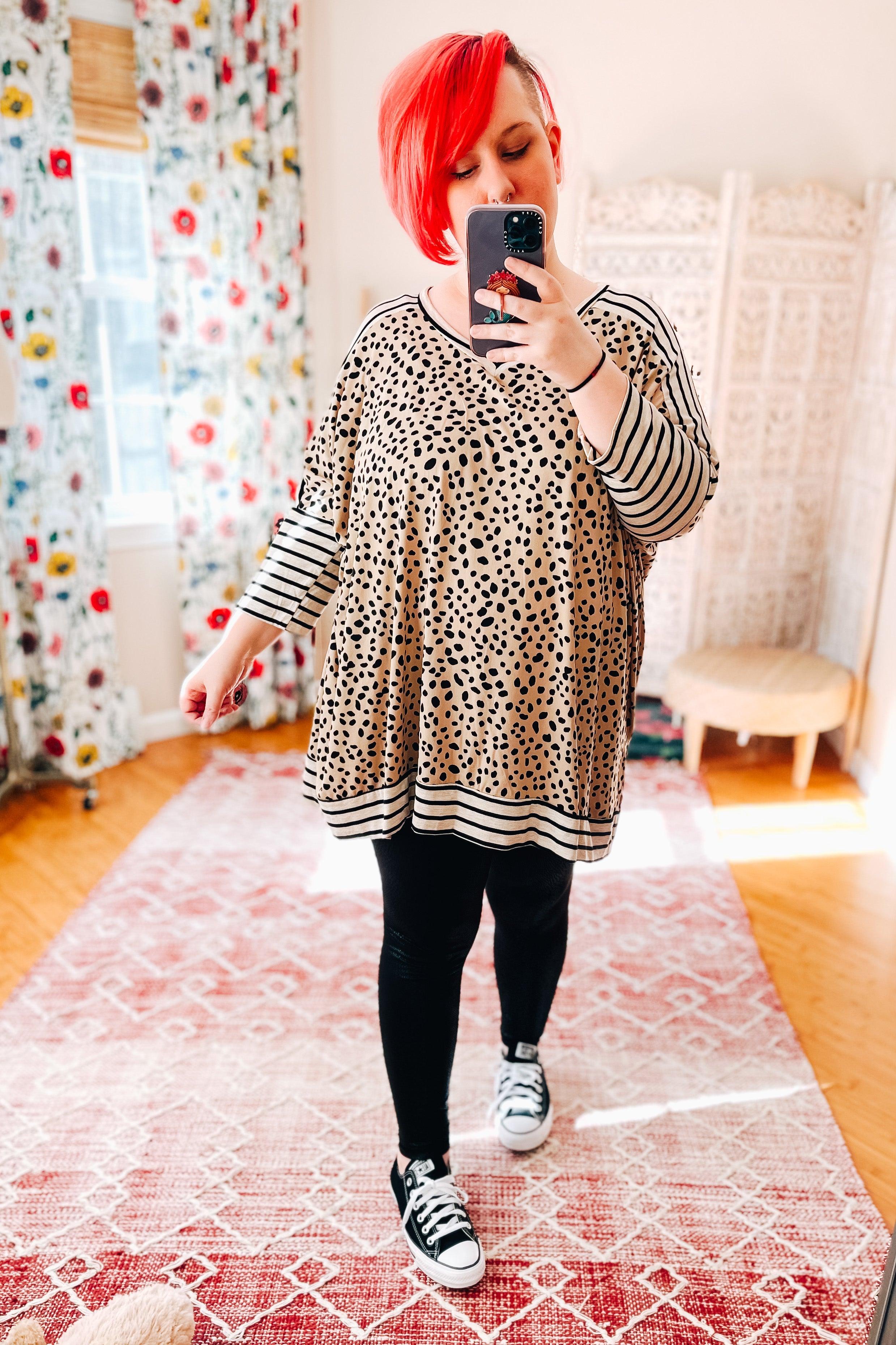 Woman taking a mirror selfie wearing a taupe tunic with scattered black spots and contrasting black and taupe horizontal stripes on sleeves and hem, paired with black leggings and black sneakers.