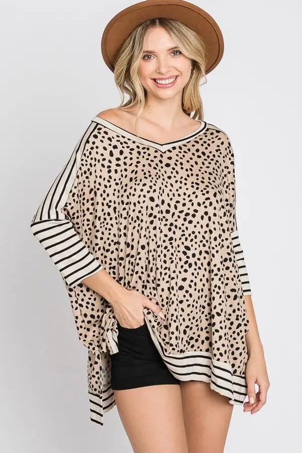 Smiling woman wearing a taupe loose-fit top with black irregular dots and contrasting black and white horizontal stripes on the sleeves and hem, paired with black shorts and a brown hat