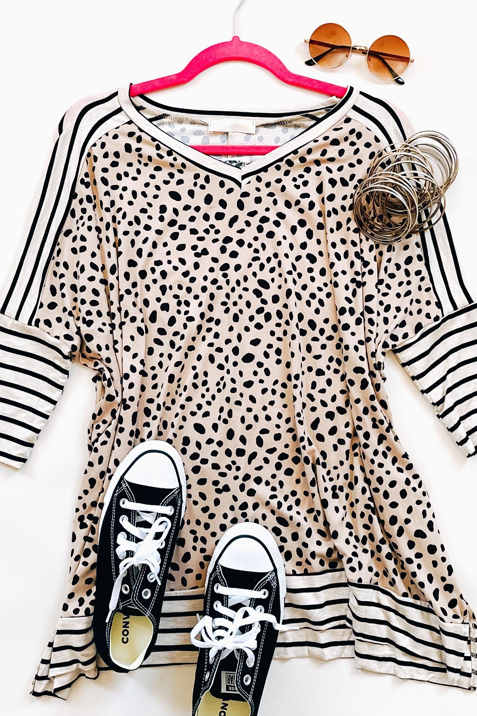 Taupe tunic with black irregular spots and contrasting black and white horizontal stripes on sleeves and hem, paired with black and white sneakers, round sunglasses, and silver bangles