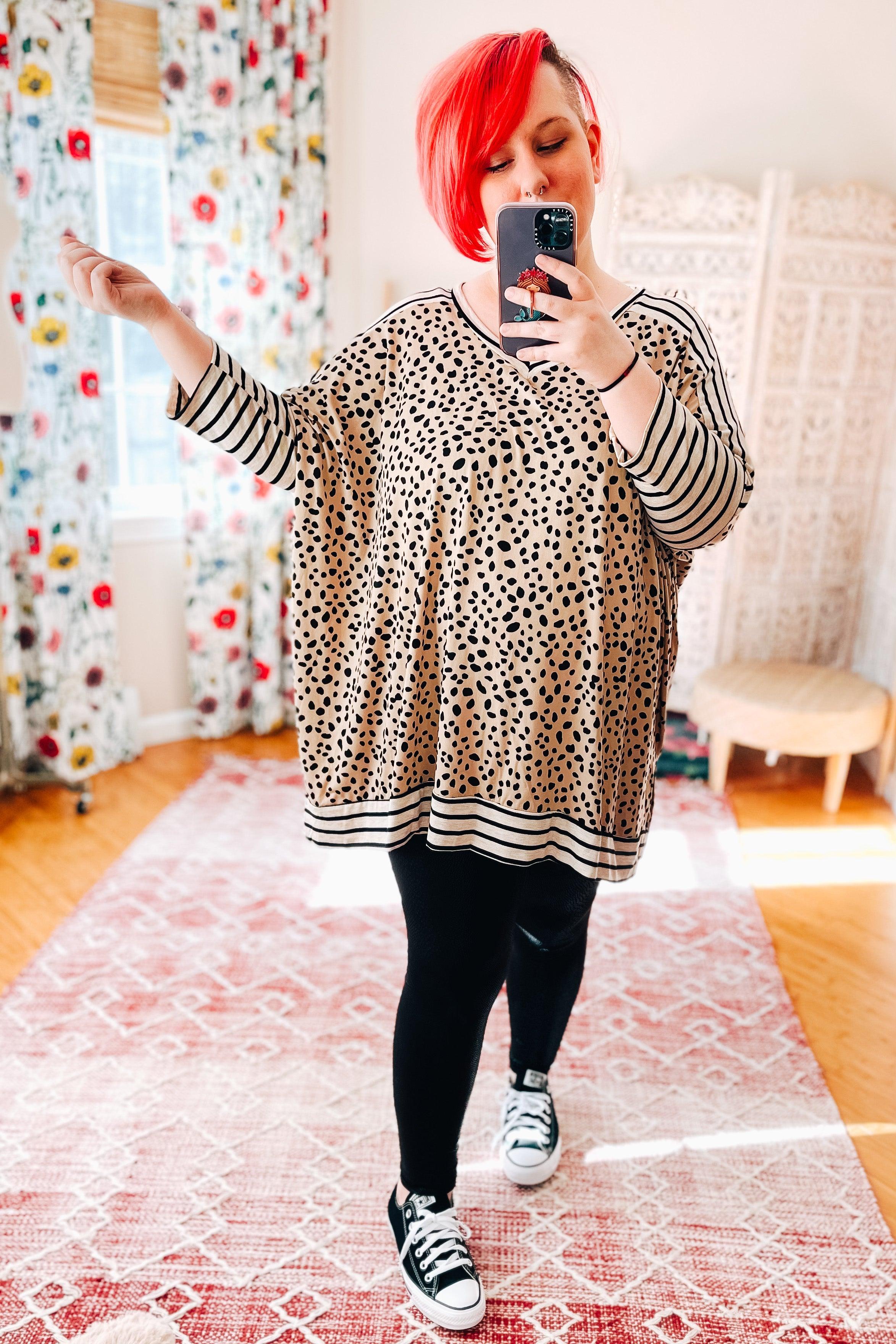 Woman taking a mirror selfie wearing a taupe loose tunic with small black spots and striped black and taupe sleeves and hem, paired with black leggings and black sneakers