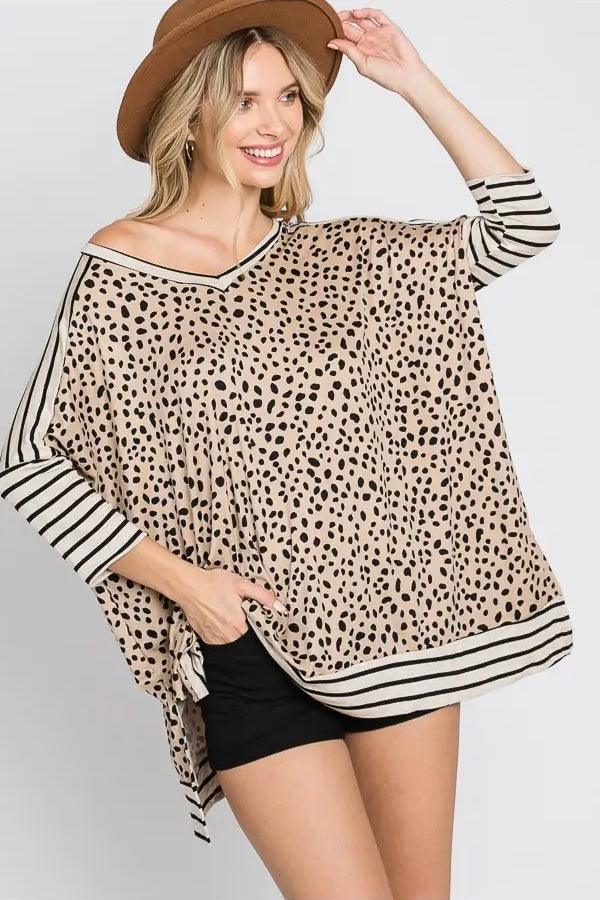 Woman wearing a taupe loose tunic with black irregular spots and black and white striped accents on sleeves and hem, paired with a brown hat and black shorts