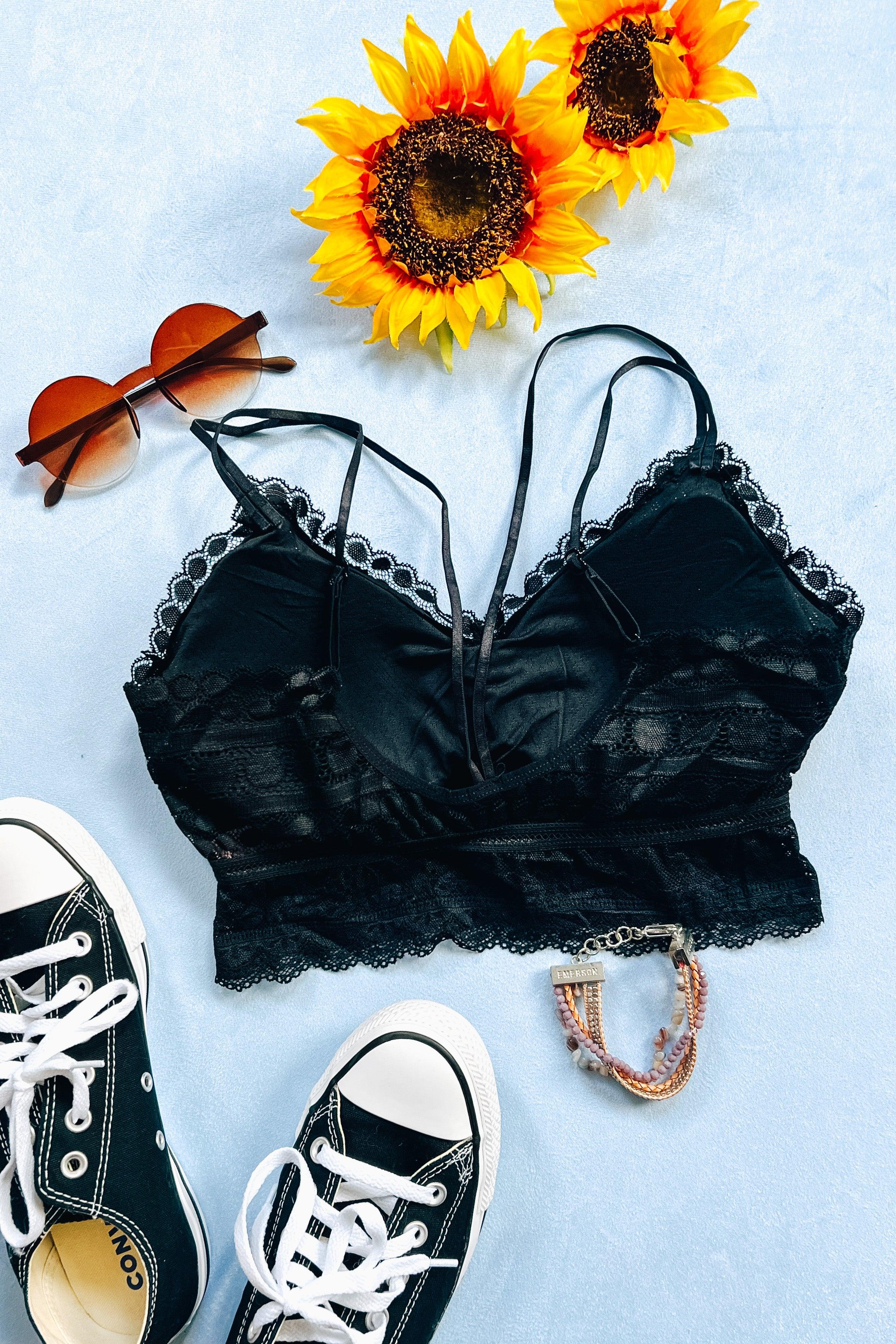 Black lace bralette with delicate scalloped edges and thin straps, styled alongside black canvas sneakers, round brown sunglasses, sunflower blooms, and a beaded bracelet on a pale blue background.