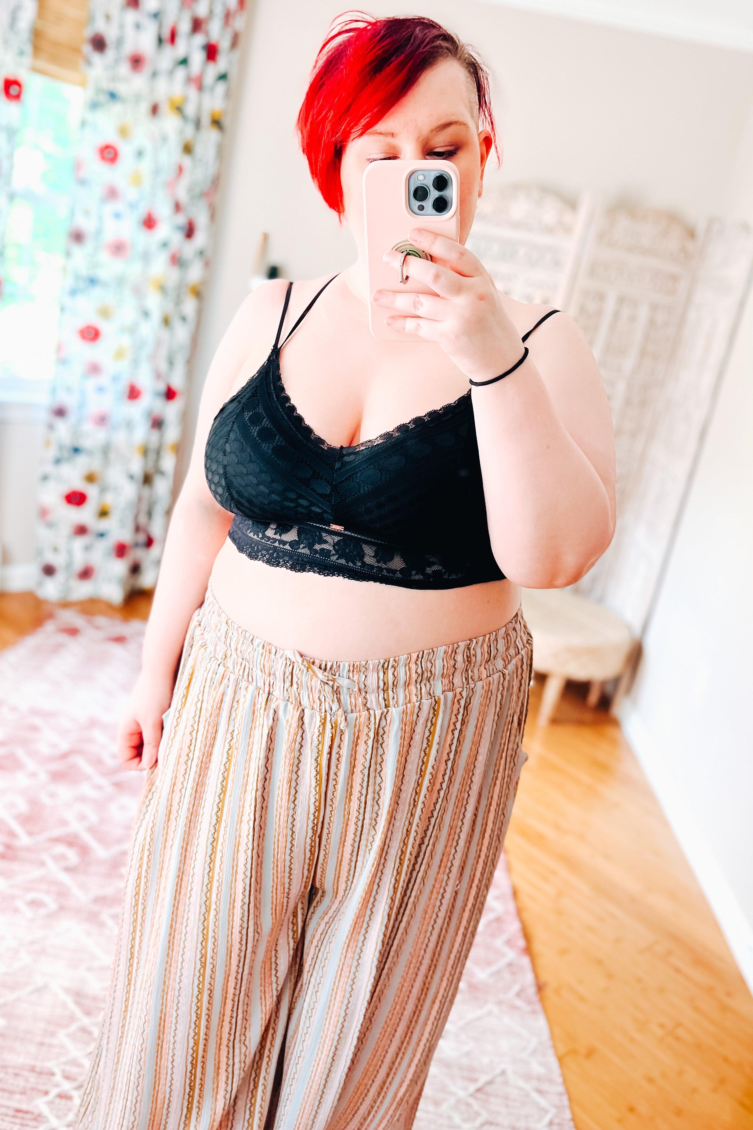 Woman with bright red hair wearing a black lace bralette with subtle geometric details and thin straps, paired with relaxed fit striped pants, taking a mirror selfie in a softly lit room.
