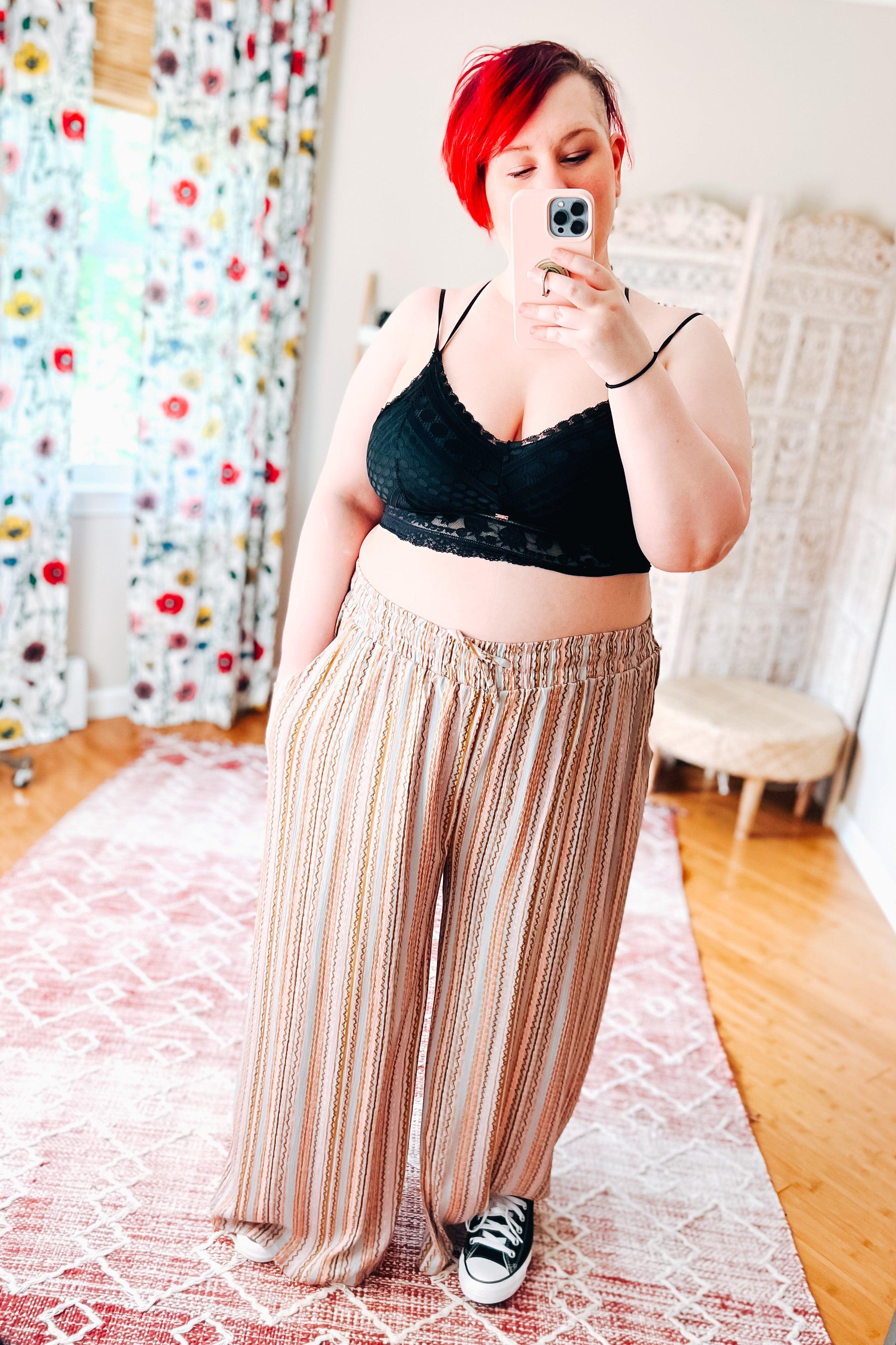 Woman with vibrant red hair wearing a black bralette with subtle texture and scalloped edge, paired with loose, striped wide-leg pants and black sneakers, taking a mirror selfie in a bright room with floral curtains.