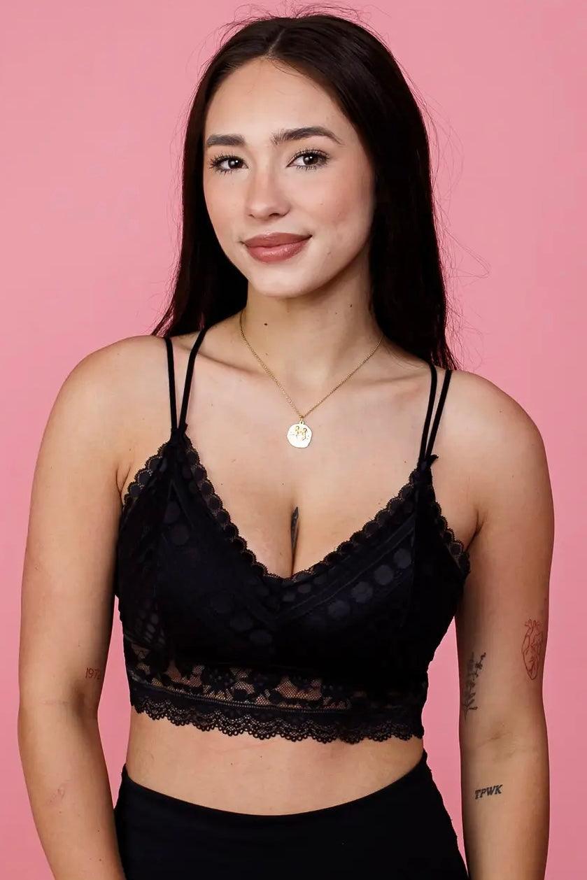 Young woman wearing a black lace bralette with thin double straps against a pink background, showcasing a delicate and elegant design.